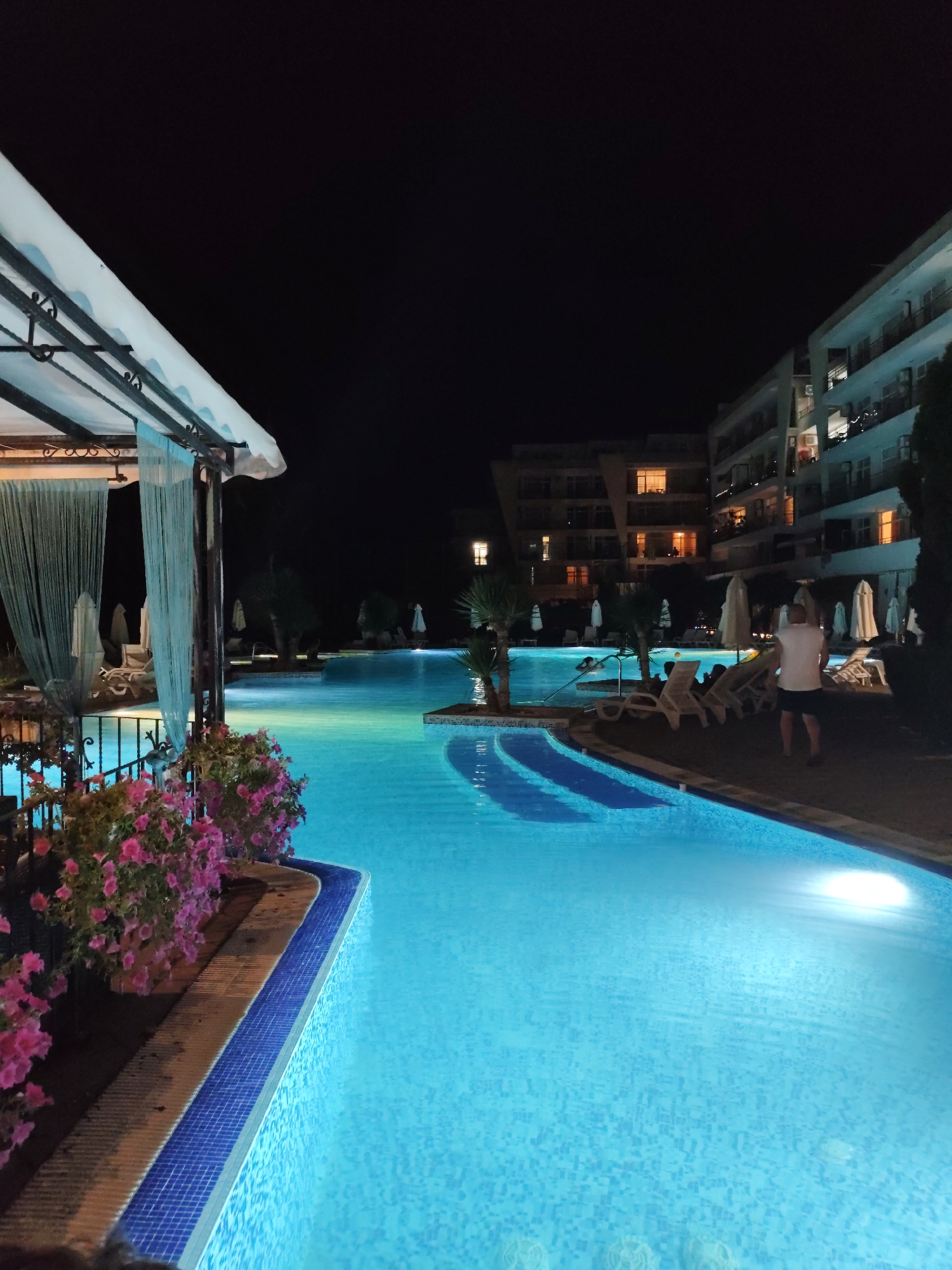 The image shows an outdoor swimming pool at night, likely at a resort or hotel in Nesebar, Bulgaria. The pool is illuminated with bright blue underwater lights, creating a vibrant glow. In the foreground on the left, there's a covered seating area with sheer curtains and pink flowers blooming along the edge. In the background, modern multi-story buildings with illuminated windows are visible, suggesting a built-up area. Several lounge chairs and umbrellas are arranged around the pool, some unoccupied. A man in a white t-shirt and dark shorts is standing near the edge of the pool, facing away from the camera. A few other people are visible in the water, swimming. The sky is dark, indicating nighttime.