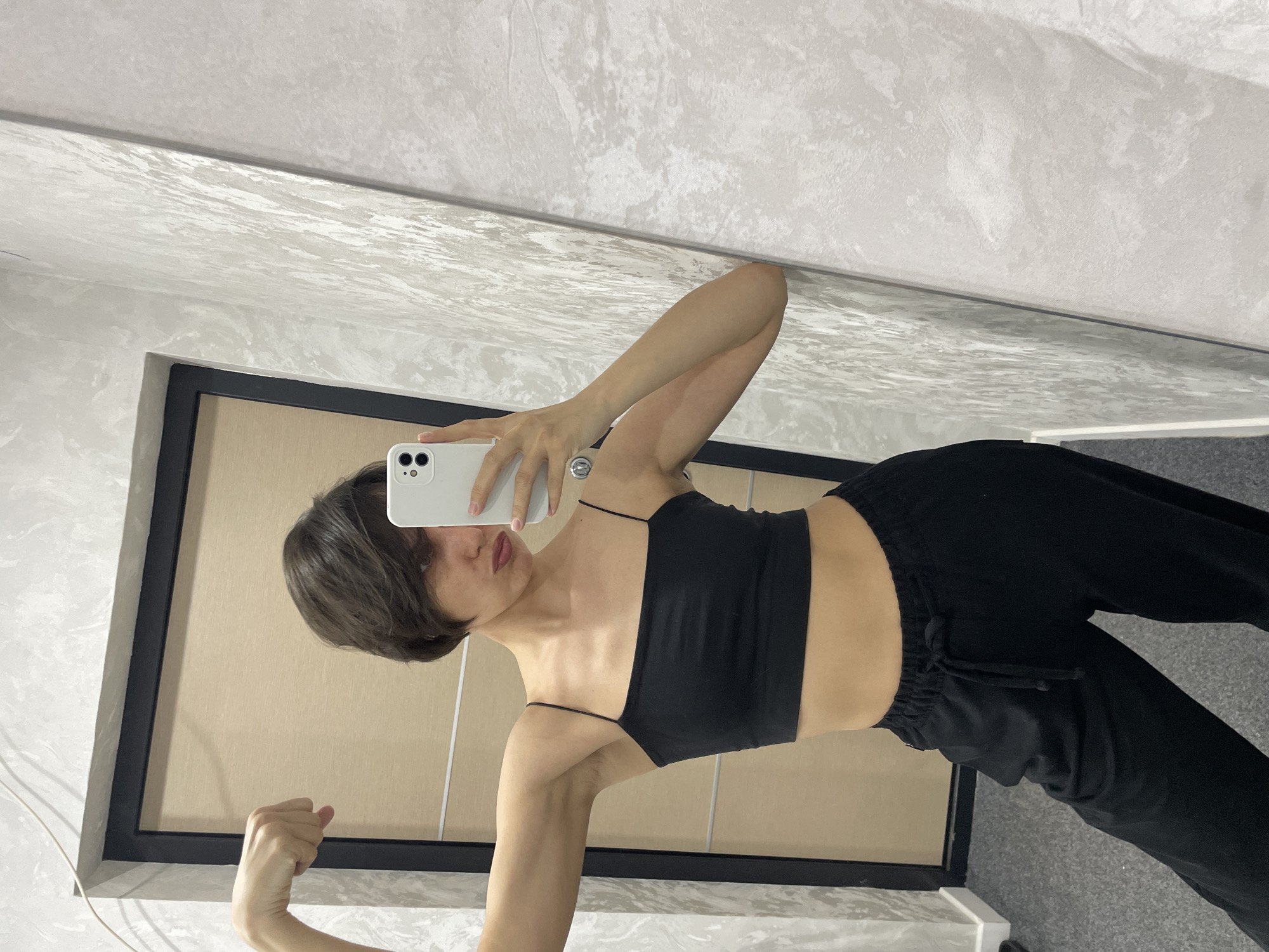 A young person with short brown hair is taking a selfie in front of a mirror. The person is wearing a black spaghetti strap crop top and black sweatpants. Their left arm is flexed. The background includes a door with a black frame and a textured wall. There is no indication of this image being from Izhevsk, Russia.