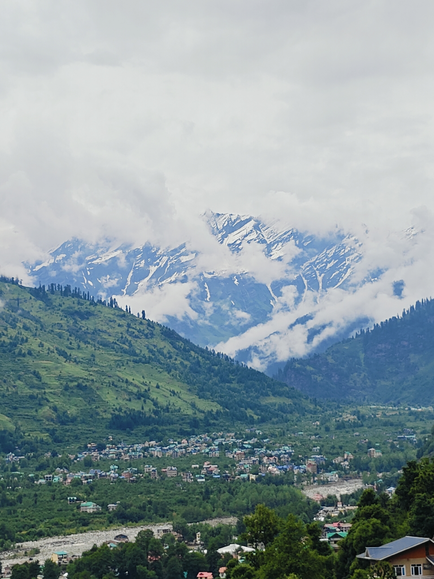 This is a photograph of a mountain village in Vashisht, India. The scene is dominated by lush green mountains that transition into snow-capped peaks in the distance. The sky is overcast with a thick layer of clouds, obscuring some of the upper portions of the mountains and lending a diffused light to the scene, suggesting it might be a cloudy day, possibly morning or late afternoon.
In the foreground and mid-ground, the mountainsides are covered in dense green vegetation, with scattered trees and some terraced fields visible. A village is nestled in the valley below, characterized by numerous small buildings with colorful roofs, clustered together amongst trees and winding roads. A riverbed, appearing dry or with minimal water, cuts through the valley floor. The architecture of the village buildings is typical of the region, with many houses featuring pitched roofs.
The overall mood is serene and picturesque, highlighting the natural beauty of the mountainous landscape and the humble human settlement within it. There is no visible text in the image. The photograph captures a wide vista, with the viewer's perspective looking down upon the village from a higher vantage point on the mountainside. The activity depicted is simply the existence of the village and its integration with the natural environment, without any specific human activity being clearly discernible due to the distance and scale.