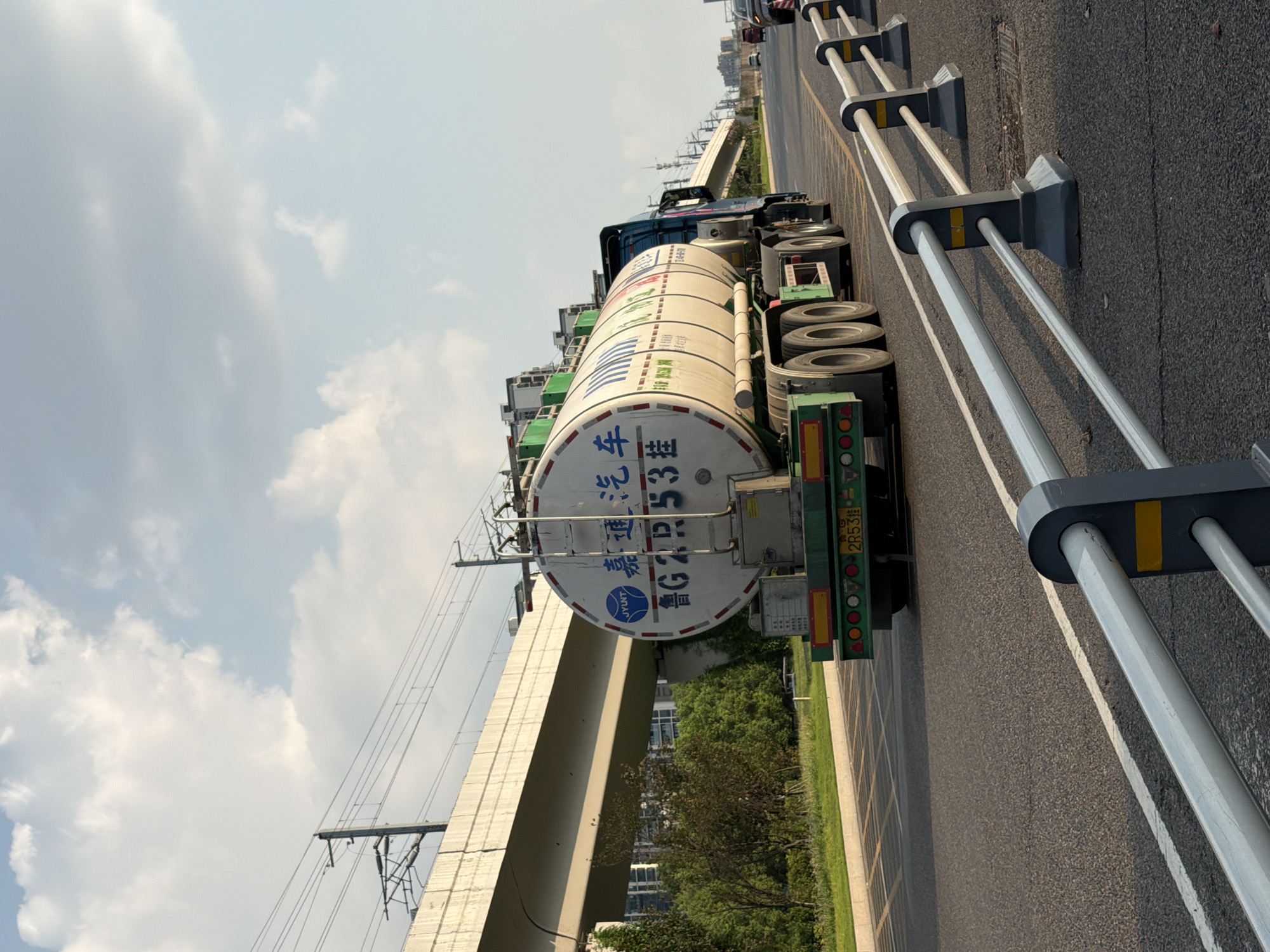 A semi-trailer truck with a white tanker trailer drives on a road in Jiangjin, China. The truck is green and blue, and the tanker is white with blue lettering. The lettering on the tanker appears to be Chinese characters. The truck is in motion, and the wheels are visible. The road is asphalt and has white lane markings. In the background, there is an elevated railway line with overhead power lines and some buildings. Trees and grass are visible on the side of the road. The sky is blue with some white clouds. The scene appears to be a sunny day.