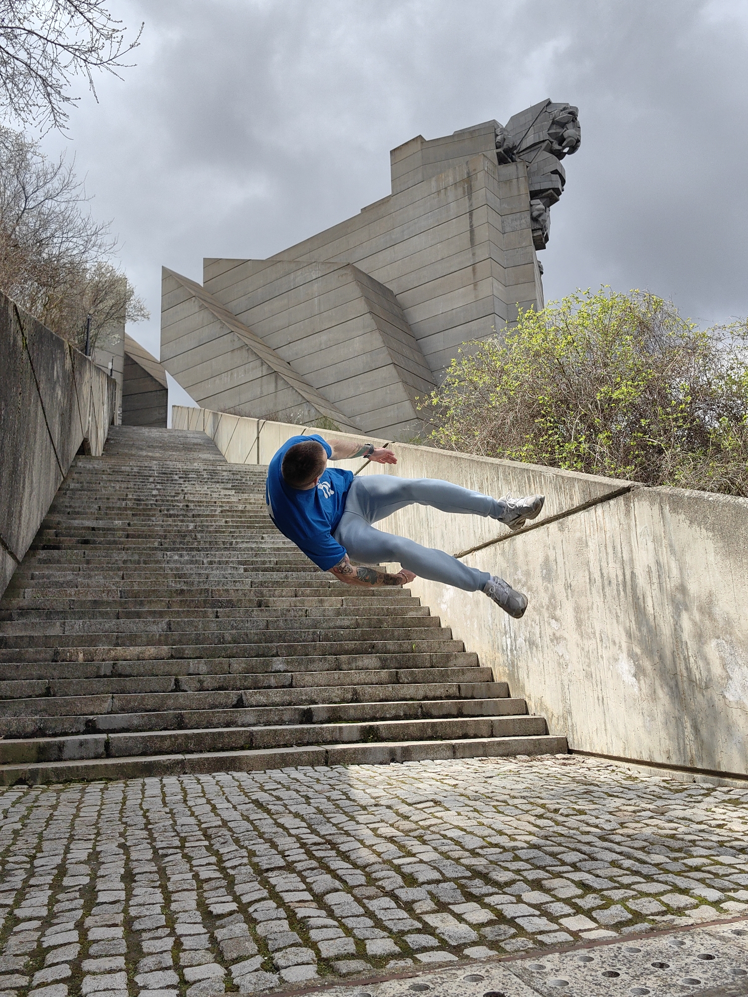 The image shows a man in mid-air, positioned diagonally across a flight of stone steps. He wears a blue t-shirt, light gray pants, and gray sneakers. His body is oriented as if he is falling or jumping. A concrete wall runs along one side of the staircase. Behind the staircase, there is a large, modernist concrete structure, with visible sculpted faces at its top, emerging above some greenery. The area below the staircase is paved with cobblestones. The sky is overcast. The location is the Monument to 1300 Years of Bulgaria in Shumen, Bulgaria.