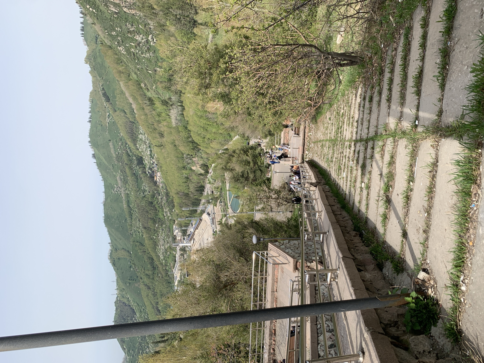 This image depicts a view from Kok-Tobe Hill in Almaty, Kazakhstan. The foreground shows a stone staircase with metal railings, descending towards a lower viewpoint. There are people gathered at the lower viewpoint. The background reveals a city view with lush greenery and mountains. The day is bright, and the scene appears calm.