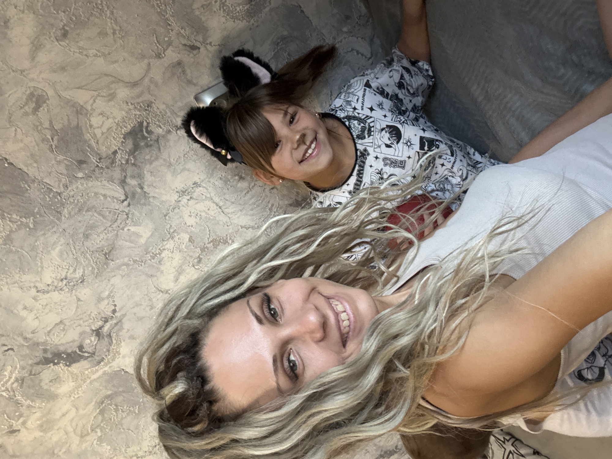 The image shows two females, an adult and a child, posing for a selfie indoors. The adult, in the foreground, has long blonde hair and is wearing a light-colored tank top. The child, in the background, has brown hair and is wearing cat ears and a t-shirt with a graphic print. They appear to be on a couch, with a textured wall behind them. The scene is consistent with a domestic setting in a residential area. There is no indication of the specific location in the city of Rusanovo, Russia.
