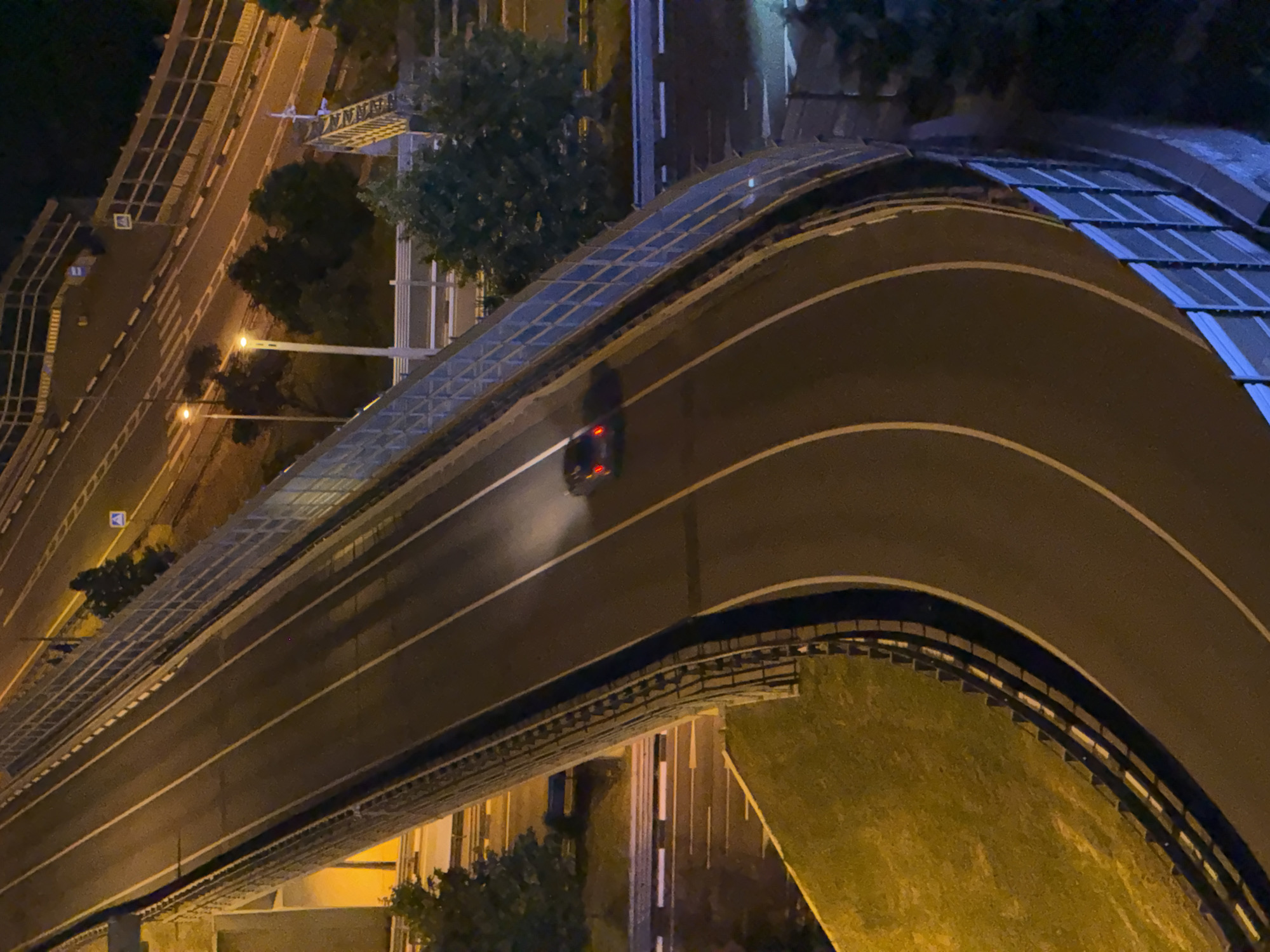 The image shows a highway overpass in the city of Sochi, Russia, at night. A dark-colored car is traveling on the overpass, emitting a plume of white smoke from its rear. The highway has multiple lanes marked with white lines. The overpass is lined with a blue-tinted sound barrier fence. Surrounding the overpass are trees and grassy areas illuminated by streetlights. In the background, other roads and structures are visible, also lit by artificial lights. The overall scene suggests an urban or suburban environment at night.