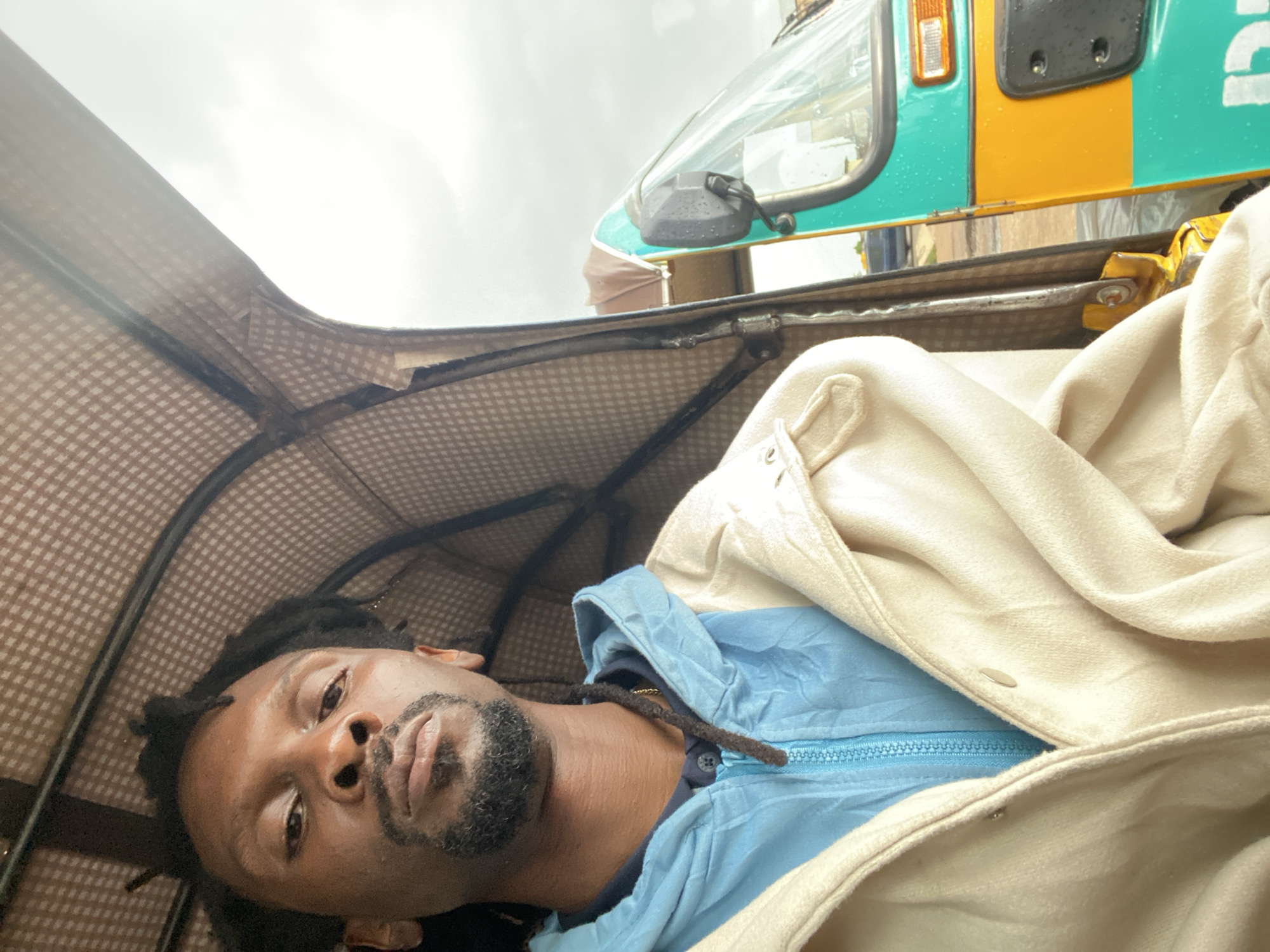The image shows a man sitting in the driver's seat of a three-wheeled auto-rickshaw, commonly known as a Keke Napep, in Gura Top, Nigeria. The man has dark skin, dreadlocks, and a goatee. He is wearing a light blue hooded jacket over a dark shirt, and a light-colored jacket on top of that. The Keke Napep has a brown checkered canopy overhead and is painted teal and yellow. The exterior of the Keke Napep is visible to the right, with a side mirror and part of the windshield. The background appears to be overcast and possibly rural, with some vegetation visible in the distance. The image is taken from a slightly upward angle, looking towards the man and the front of the vehicle.