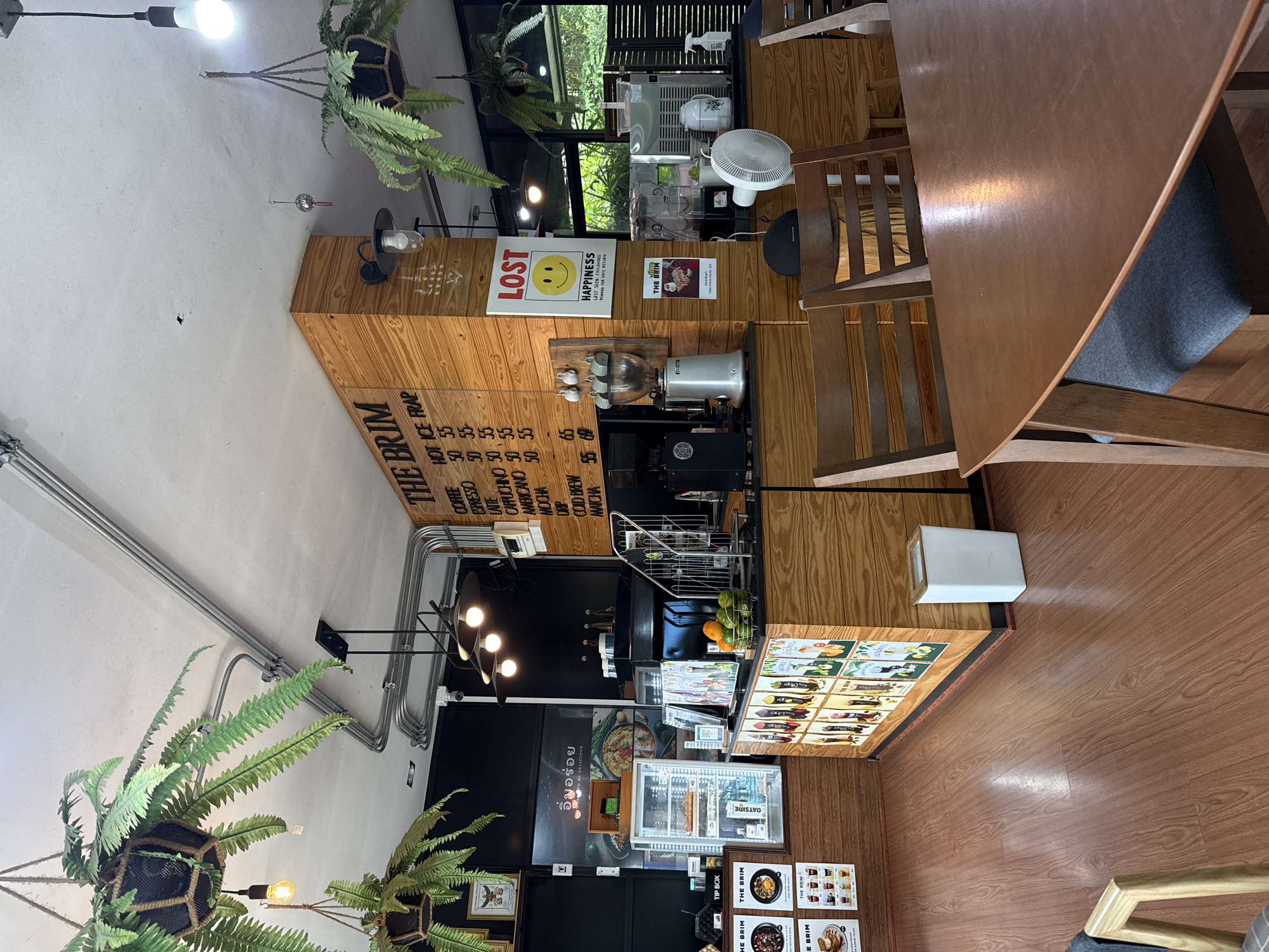 The image shows the interior of "The Brim" cafe, located in Tha Chana, Thailand. The cafe has a rustic and modern design, with exposed pipes, wooden accents, and hanging plants. The scene is captured from a perspective that includes a dining table in the foreground, with wooden chairs.
In the background, a counter area is visible, featuring a coffee grinder, espresso machine, and a refrigerator displaying food items. A menu board with prices for various coffee drinks is displayed prominently on a wooden pillar. A sign with a smiley face and the word "LOST" is also visible. There are no people in the image. The environment appears clean and organized, suggesting a well-maintained establishment.