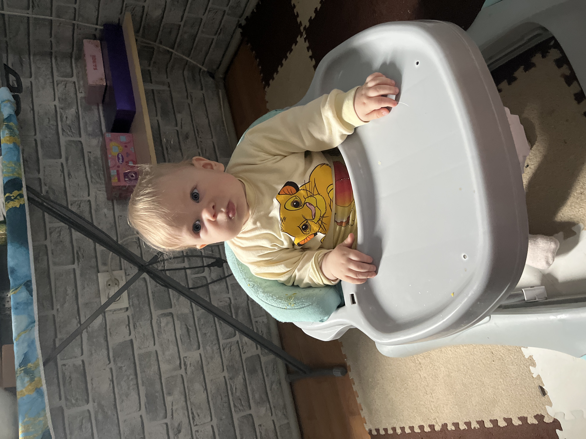 A young child with blond hair is seated in a gray highchair, wearing a yellow long-sleeved shirt with a lion cartoon character on it. The child is looking directly at the camera. The highchair tray has some food debris on it. The background shows a wall with gray brick-patterned wallpaper, and a floor covered with interlocking foam mats of varying colors. A shelf with some boxes is visible on the wall. The image was taken indoors. The location in the city of Vesyoloye, Russia is unknown.