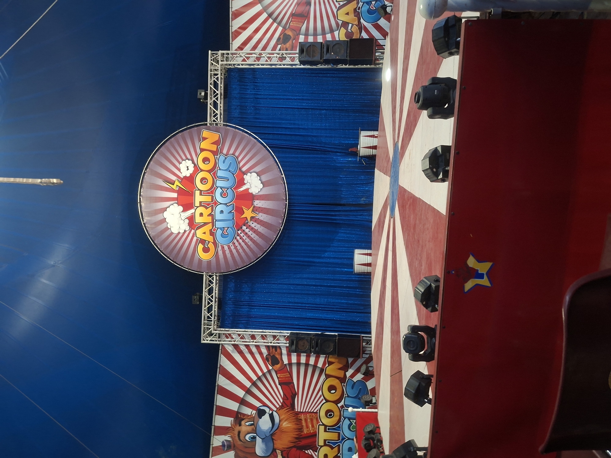 The image shows a circus tent interior, likely in Unknown, Unknown. A large circular sign reads "CARTOON CIRCUS" with cartoonish fonts and illustrations. The sign is mounted on a metal truss structure in front of blue fabric curtains. Above the sign, there are speaker boxes. Below the sign, another panel with a cartoon animal character is visible, also advertising the circus. To the right, a red and white striped structure is partially seen, equipped with multiple lighting fixtures and a yellow and blue star logo. A chair is visible to the right, suggesting seating for the audience. The overall setting is colorful and designed to be visually appealing for a circus performance.