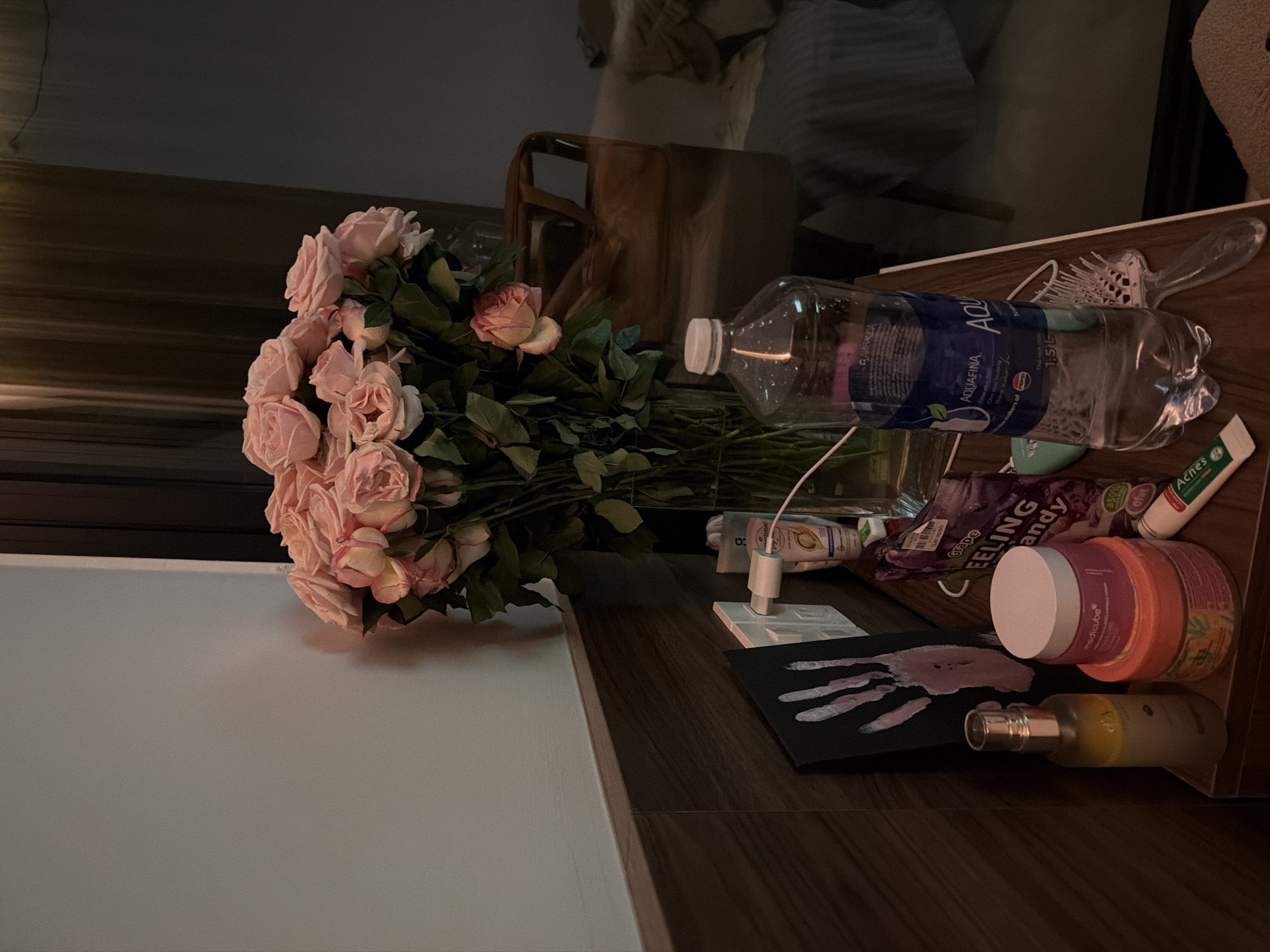 The image depicts a bedside table in a room in Đà Nẵng, Vietnam. On the table, a bouquet of pink roses is displayed in a clear vase. Next to the vase, an Aquafina water bottle stands upright, with condensation visible on its surface. To the left of the bottle, several cosmetic products are arranged, including a bottle with a silver spray top, two jars, and a tube labeled "Acnes". A purple packet labeled "FEELING candy" is also present. A small black canvas with a pink handprint is leaning against the wall, behind the cosmetic items. The wall behind the table is painted a light gray and is partially covered by wooden paneling at the bottom. A white electrical outlet with a plug inserted is visible on the wall. In the background, a wooden chair and part of a bed can be seen, suggesting a bedroom setting. The lighting in the room is dim, casting soft shadows.