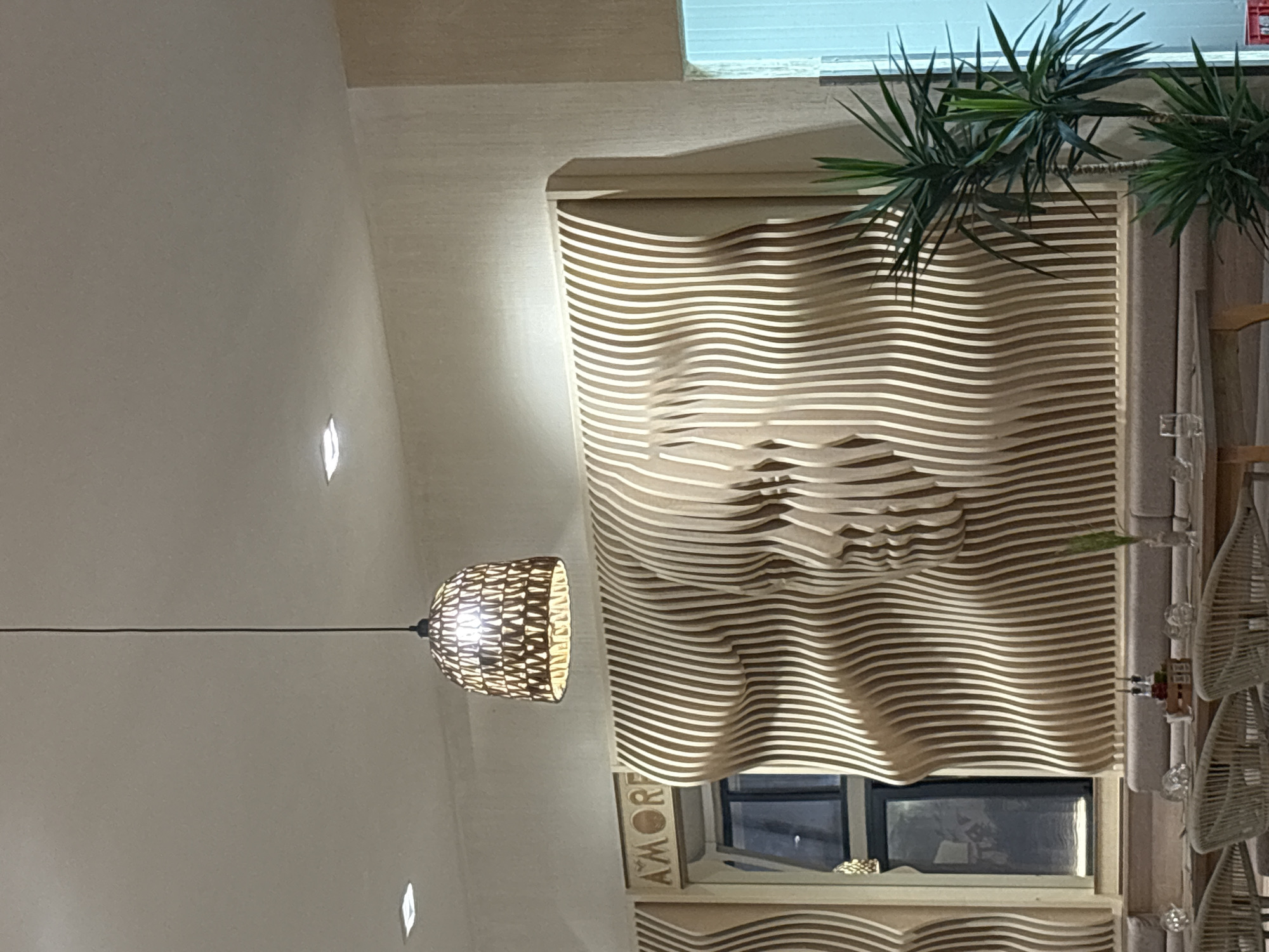 The image shows the interior of a restaurant with a distinctive wall decoration. The wall is made of wooden slats arranged in a wavy pattern, creating an abstract representation of two faces. Above the wall art, the word "AMORE" is visible in raised lettering. A modern pendant light hangs from the ceiling, casting a warm glow. In the foreground, a long upholstered bench runs along the wall, with tables and chairs set up for dining. A tall green plant is positioned to the right of the wall art. The overall ambiance appears to be sophisticated and contemporary.