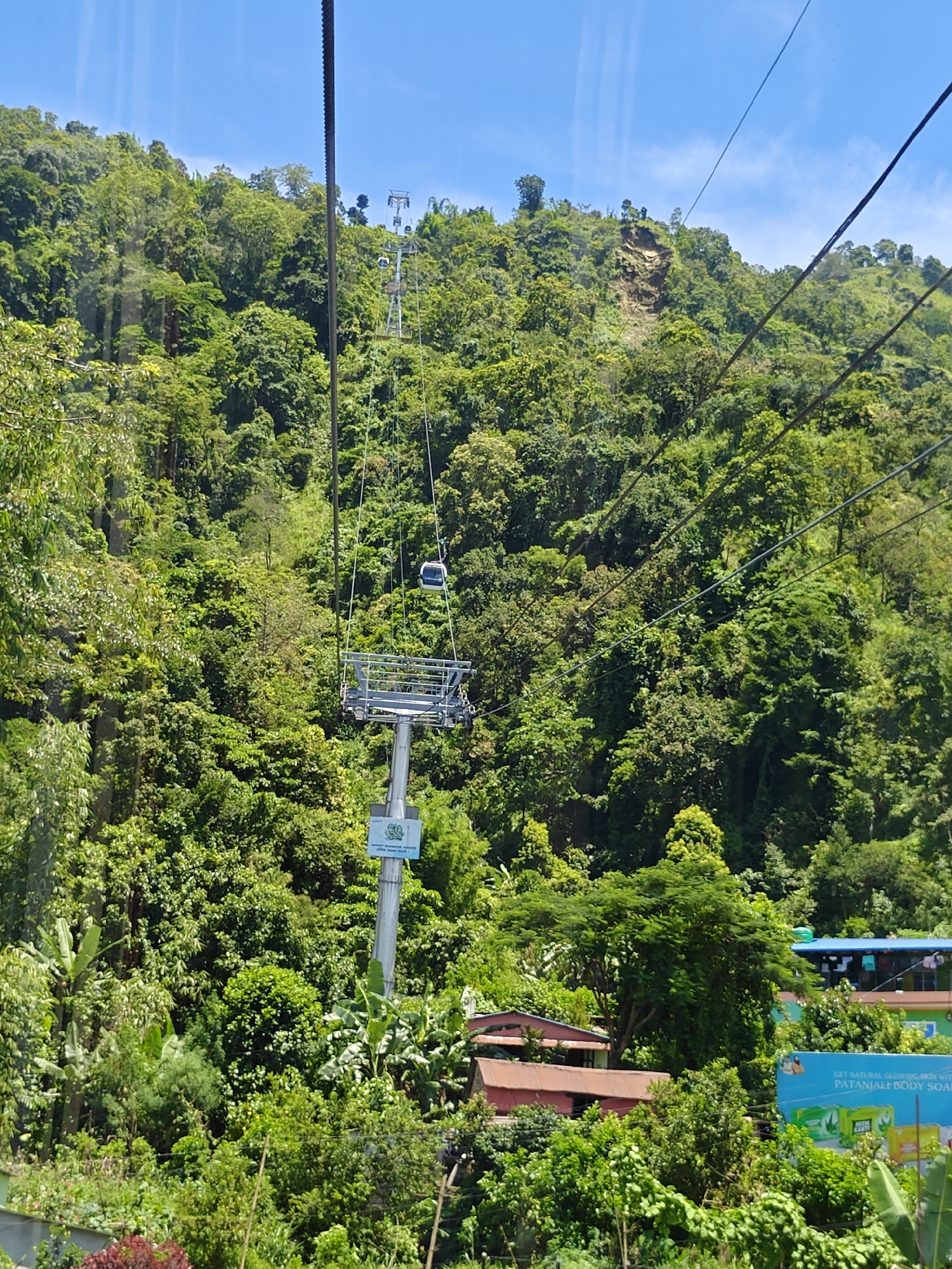 The image shows a cable car traversing a lush, green, forested mountainside in Pokhara, Nepal. The cable car is suspended from thick cables, with one car visible in the upper center of the frame. Below the cable car, a tower supports the cables, with a sign attached to its base. The mountainside is densely covered with trees and vegetation, suggesting a natural and somewhat remote environment. The sky is clear and blue, indicating good weather. In the lower portion of the image, there are glimpses of buildings, possibly homes or businesses, nestled amongst the trees, indicating some human settlement within the natural landscape. The scene evokes a sense of travel and exploration, with the cable car offering a means to ascend the steep terrain.
