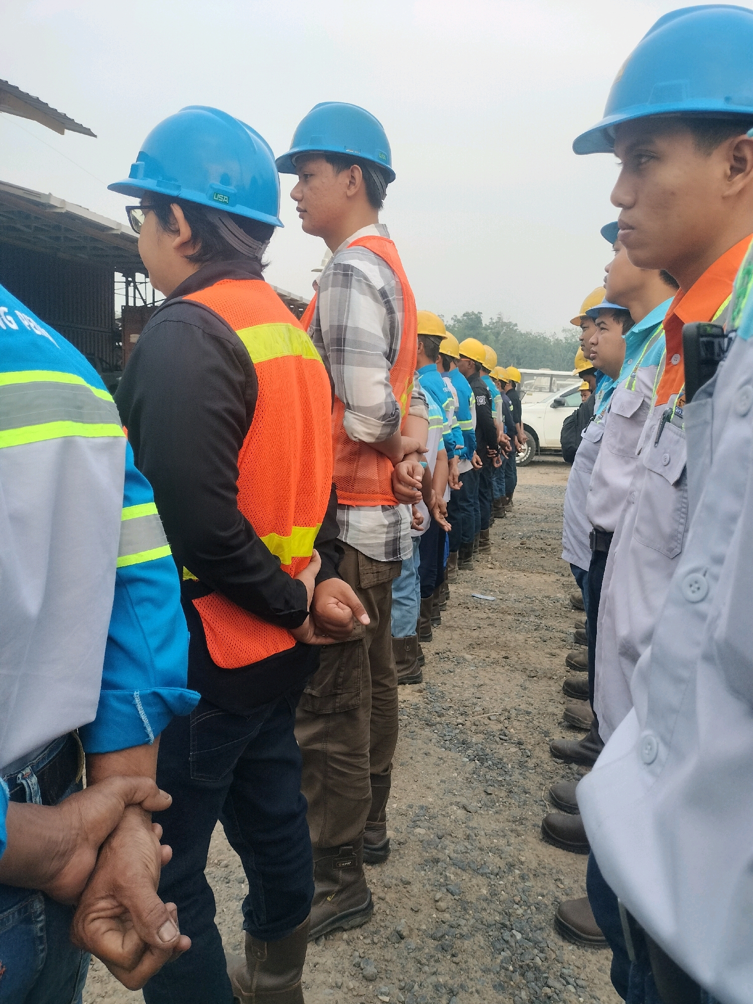 In Maburai, Indonesia, a group of male construction workers, all wearing hard hats and safety vests, stand in a line outdoors. The workers are dressed in various work attire, including shirts, long pants, and boots. Several of the workers have their hands clasped behind their backs. The environment appears to be a construction site with gravel on the ground and a building structure in the background. The sky is overcast.