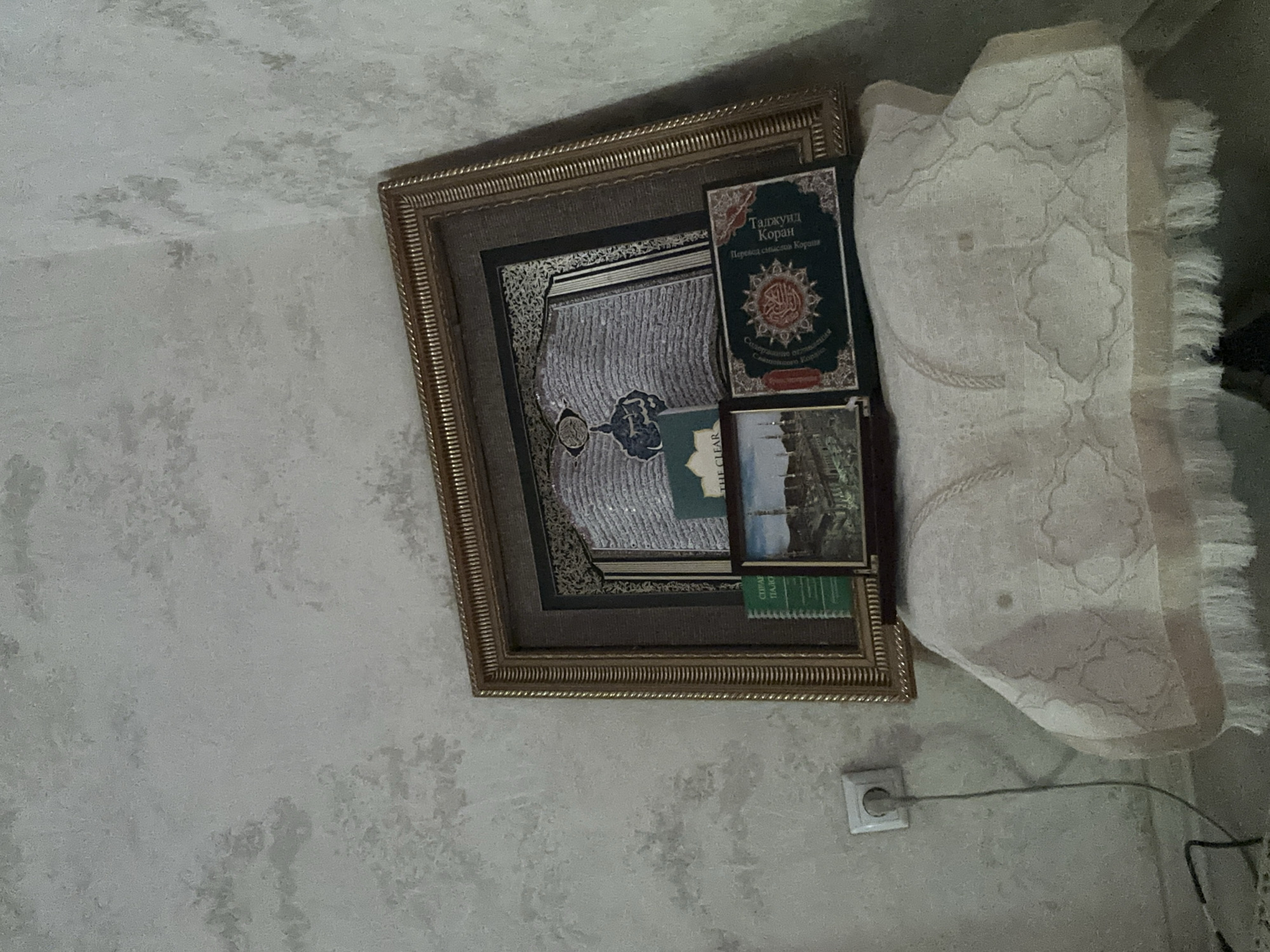 A framed artwork of an open book with Arabic script hangs on a textured, light-colored wall. In front of the artwork, on a veiled surface, are three small books. The book on the left is green with white text and a picture of a mosque. The middle book is light green and has a partially visible title. The book on the right is dark green with red and gold embellishments and Arabic script. A white electrical outlet with a plugged-in cord is visible on the wall to the left of the framed artwork. The veiled surface appears to be a draped cloth with a repeating pattern and fringe. The scene is dimly lit. This image was taken in the city of Dushanbe, Tajikistan.