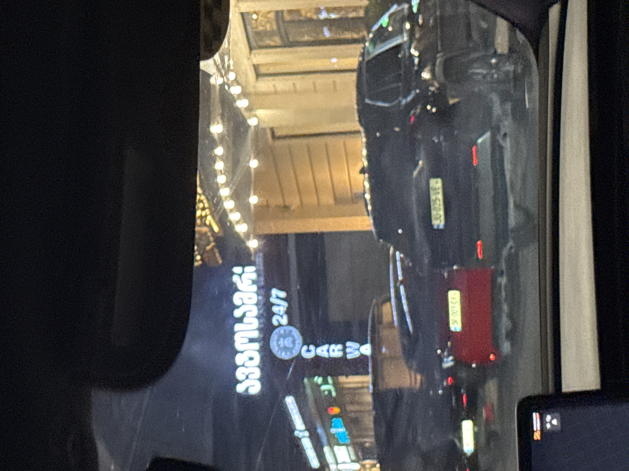 The image shows a night scene in the city of Tbilisi, Georgia, as viewed from inside a vehicle. In the background, a brightly lit sign with Georgian script, which translates to "Car Wash," is visible. Below the Georgian script, a circular emblem and the text "24/7" are displayed. Vertically arranged, the letters "CARW A" are illuminated, further indicating the establishment is a car wash. Several vehicles are present on the road, including a red car with a license plate that appears to be "DR-001-CI". The street is illuminated by streetlights and the lights from the surrounding buildings and vehicles. The overall impression is that of a typical urban night scene with an active commercial establishment.