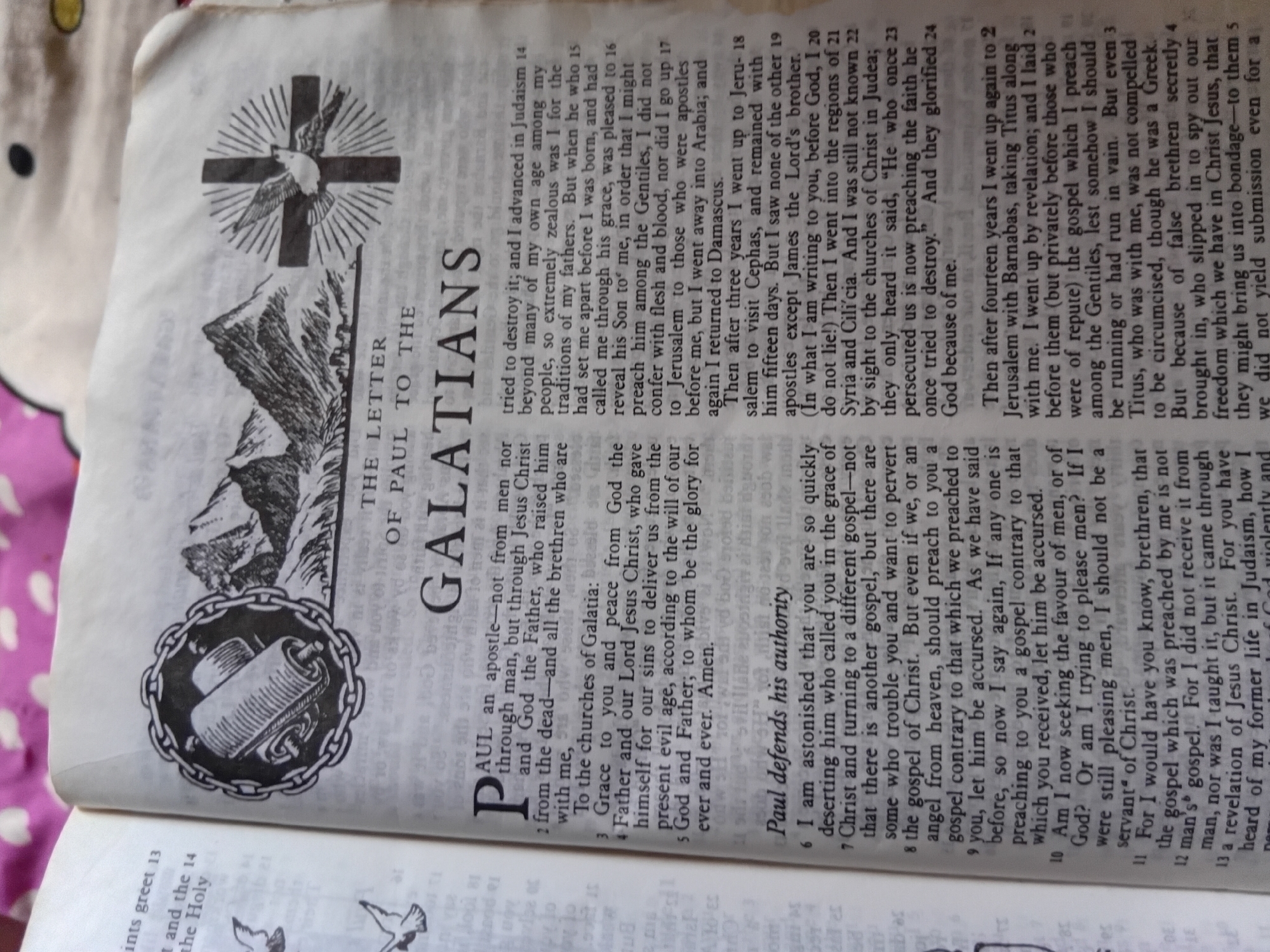 The image shows an open book, likely a Bible, with text and illustrations. The book is lying on a white fabric with a subtle pink polka dot pattern.
The primary focus is on a page displaying the text of the Epistle to the Galatians, indicated by the large heading "GALATIANS" in bold, serif font. Above the chapter title, there is a black and white illustration of a cross with radiating lines, and a bird (possibly a dove) descending towards it, set against a mountainous landscape. To the left of the main text, another illustration depicts a scroll bound by a chain.
The text itself is dense and appears to be in English. Specific verses are numbered, and headings like "Paul defends his authority" are visible. The pages are slightly yellowed, suggesting the book is old.
There are no people directly visible in the image, only the illustrations within the book. The setting appears to be indoors, given the lighting and the fabric it rests upon. The overall impression is of a religious text being studied or displayed. The provided location context of Nkpor, Nigeria, suggests this might be a Bible being used in that region.