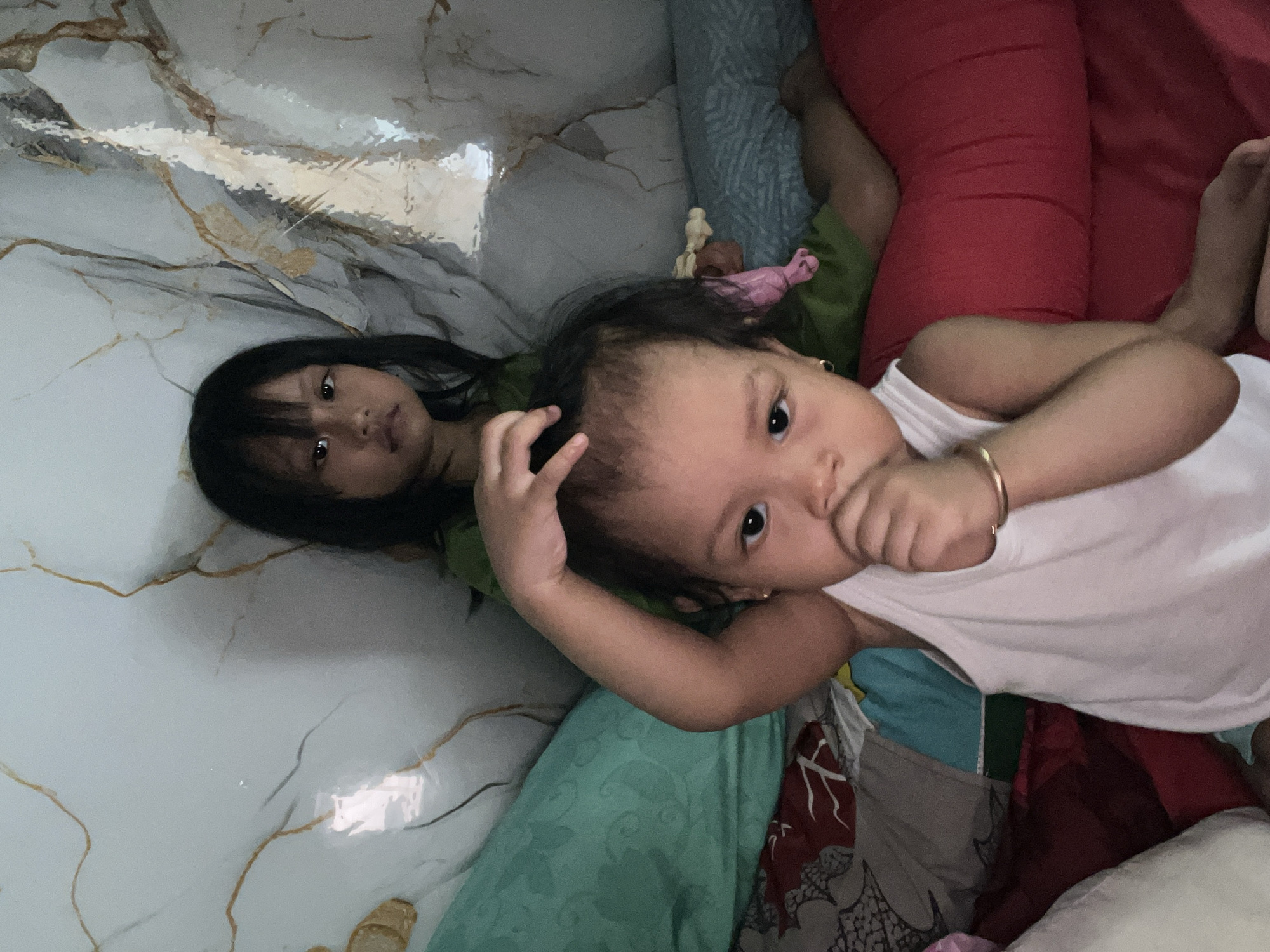 Two young girls are lying on a bed. The girl in the foreground is wearing a white tank top and has her hand to her mouth. She is wearing a gold bracelet on her left wrist. The girl in the background is wearing a green shirt and has her hand on her head. The room has patterned walls and is filled with pillows and blankets. The scene appears to be a bedroom in Kecamatan Pati, Indonesia.