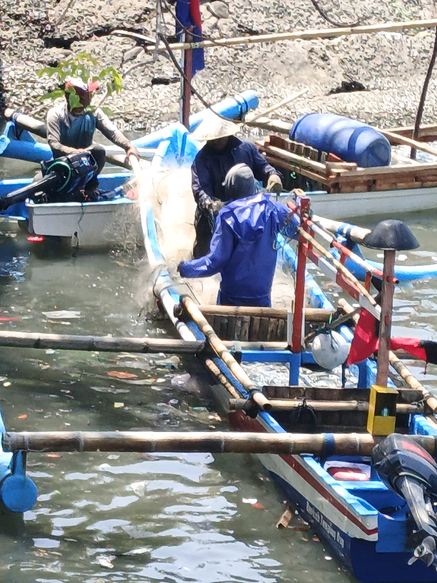 In Kelurahan Tegalkamulyan, Cilacap, Indonesia, three individuals are engaged in fishing activities on a body of water cluttered with debris. Two individuals are in a boat in the foreground, one wearing a conical hat and a blue jacket, the other in a darker garment. They are pulling a net from the water. A third individual, wearing a lighter colored shirt and a hat adorned with leaves, is seated in a boat to the left, observing the process. The boats are made of wood and bamboo, with some adorned with colorful stripes and flags. Several blue barrels and a yellow box are visible on one of the boats. The water is murky and filled with various floating objects, suggesting pollution. The background shows a bank with rocks and vegetation.