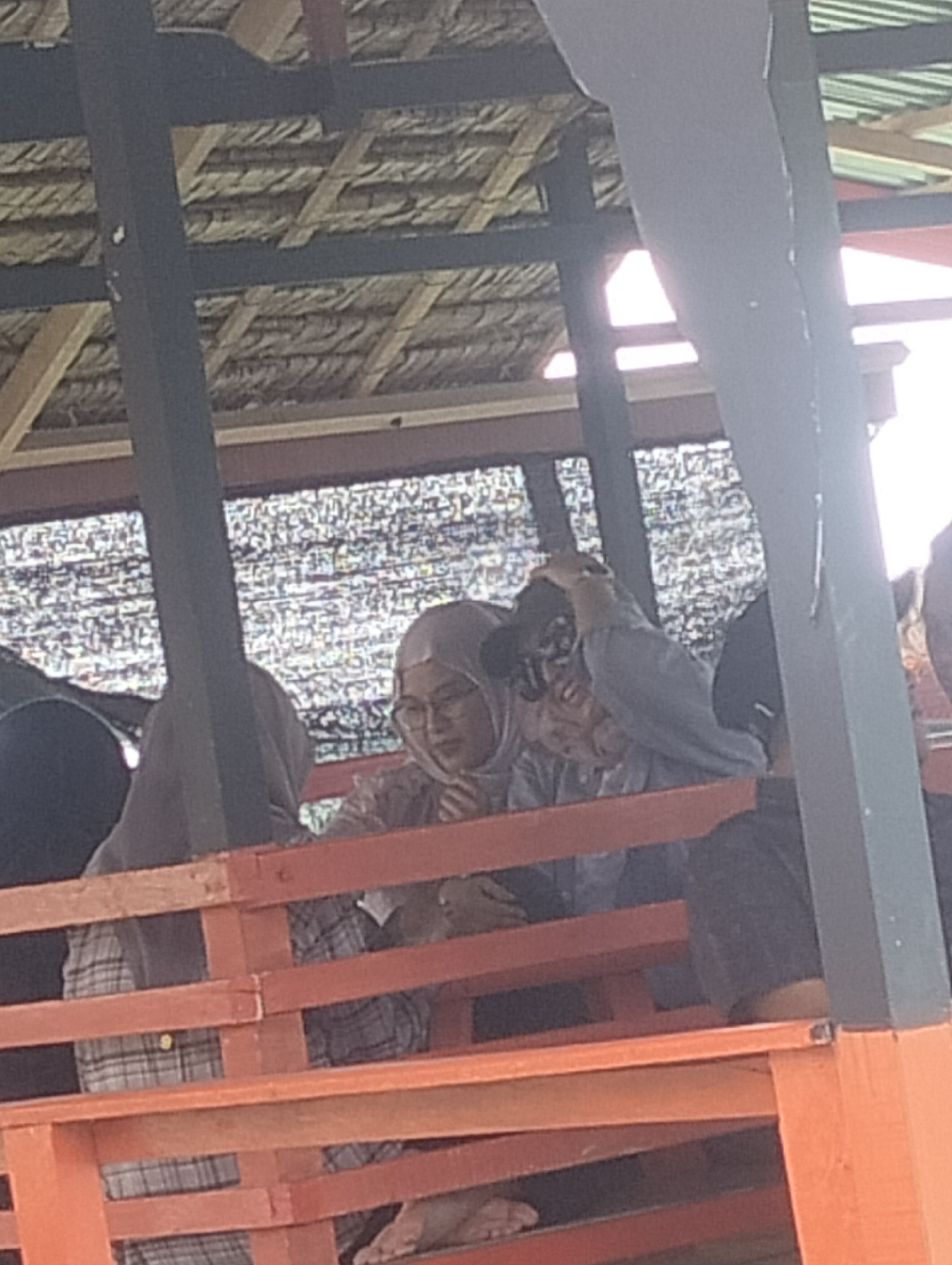 In Lancok, Indonesia, a group of people are seated on an orange wooden bench. The bench is located under a thatched roof supported by dark wooden beams and posts, creating a shaded area. The background is filled with what appears to be a patterned screen or netting.
In the foreground, a person wearing a grey plaid shirt is visible, with their legs crossed and bare feet resting on a lower wooden plank. To their right, another person is partially visible, also seated.
In the middle ground, a woman wearing a light-colored hijab and glasses is looking towards the right, with her right hand near her face. Next to her, another person, possibly a man, is wearing a dark baseball cap and a grey long-sleeved shirt, with their right hand touching the brim of their cap and their left hand in front of their face.
A person in a dark grey hijab is seated to the far left, with their back partially to the camera. Another individual wearing a dark shirt is seated to the far right, with their legs bent and feet visible. The overall scene suggests a casual gathering or waiting area.