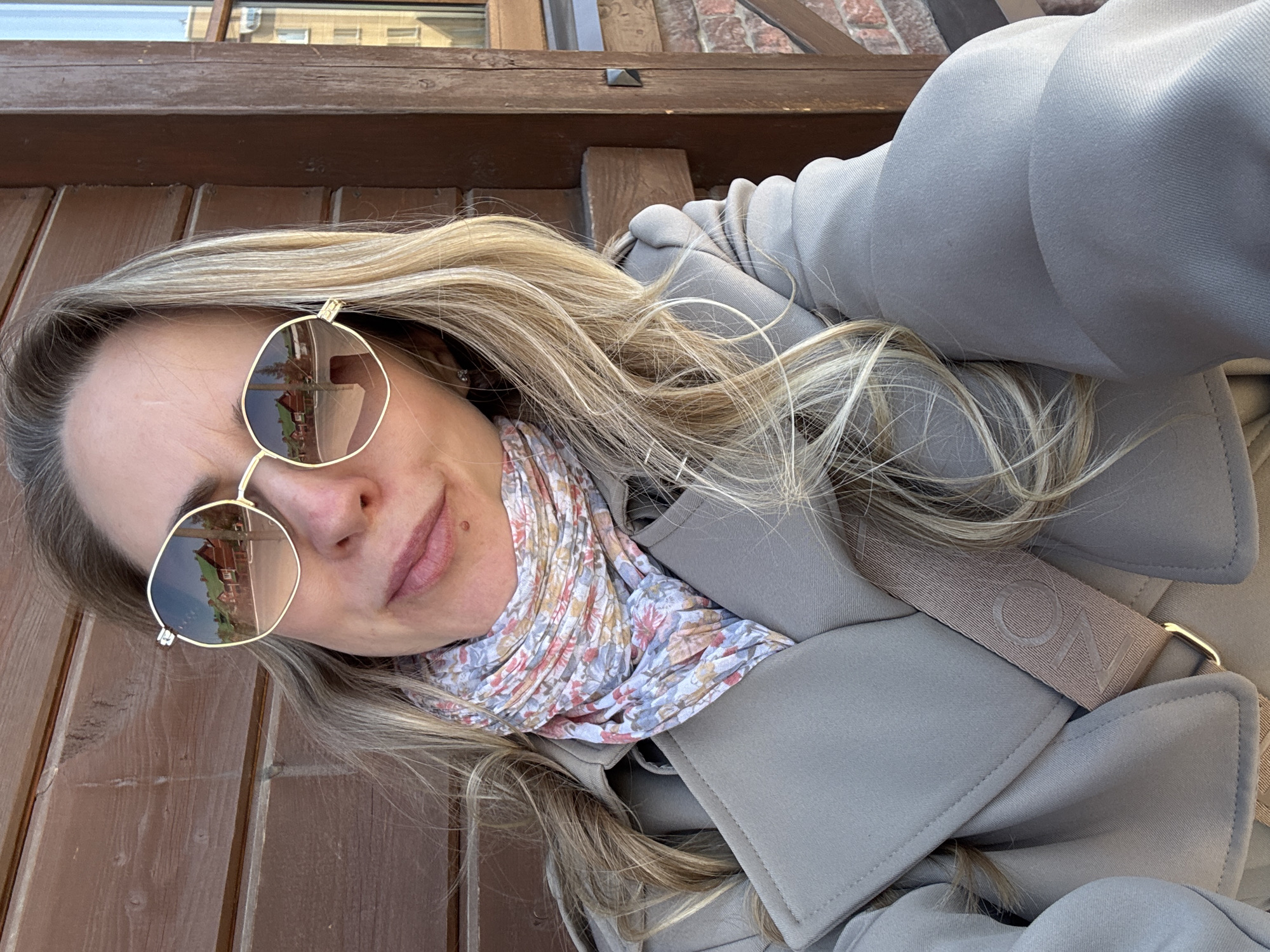 A woman is taking a selfie in the city of Chaikovsky, Russia. She is wearing a grey coat and sunglasses that reflect a building, likely a church, in the frame. She is also wearing a patterned scarf around her neck. The background consists of wooden paneling and a brick structure, possibly part of a building or outdoor seating area. The lighting indicates it is daytime.