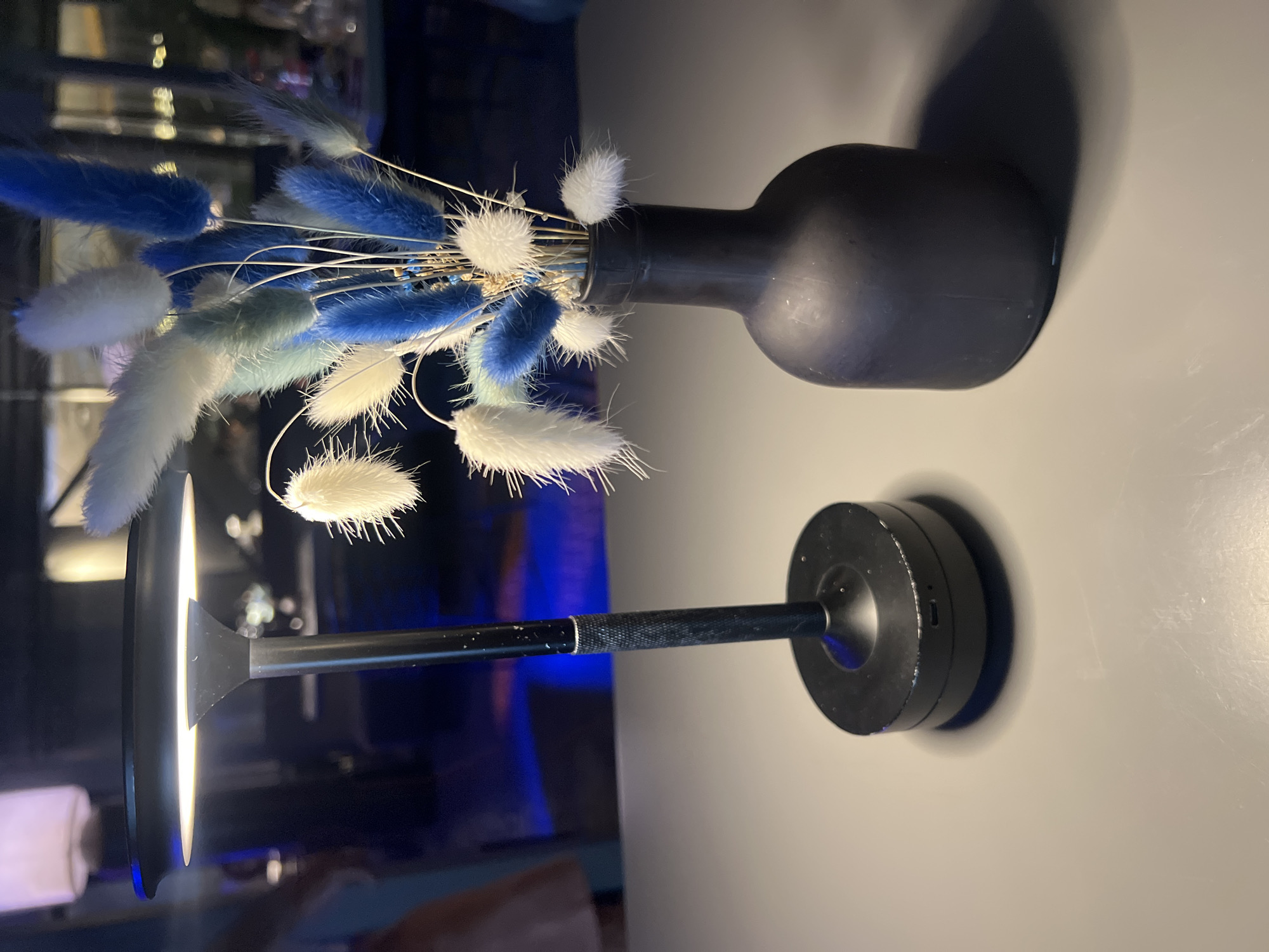 The image depicts a still life arrangement on a light gray surface. To the left, a black table lamp with a wide, flat head illuminates the scene with a warm glow. The lamp has a textured metallic stem and a circular base. To the right, a matte black vase holds a bouquet of dried bunny tail grass. The grass is a mix of white, light blue, and darker blue hues, with some green undertones visible. The background is dark and out of focus, with hints of blue lighting, suggesting an indoor setting, possibly a restaurant or a bar. No people are clearly visible in the frame, though a blurred figure is partially seen on the far left. The overall ambiance is calm and decorative. There is no indication of the city of Tiraspol, Moldova within the image or its context.