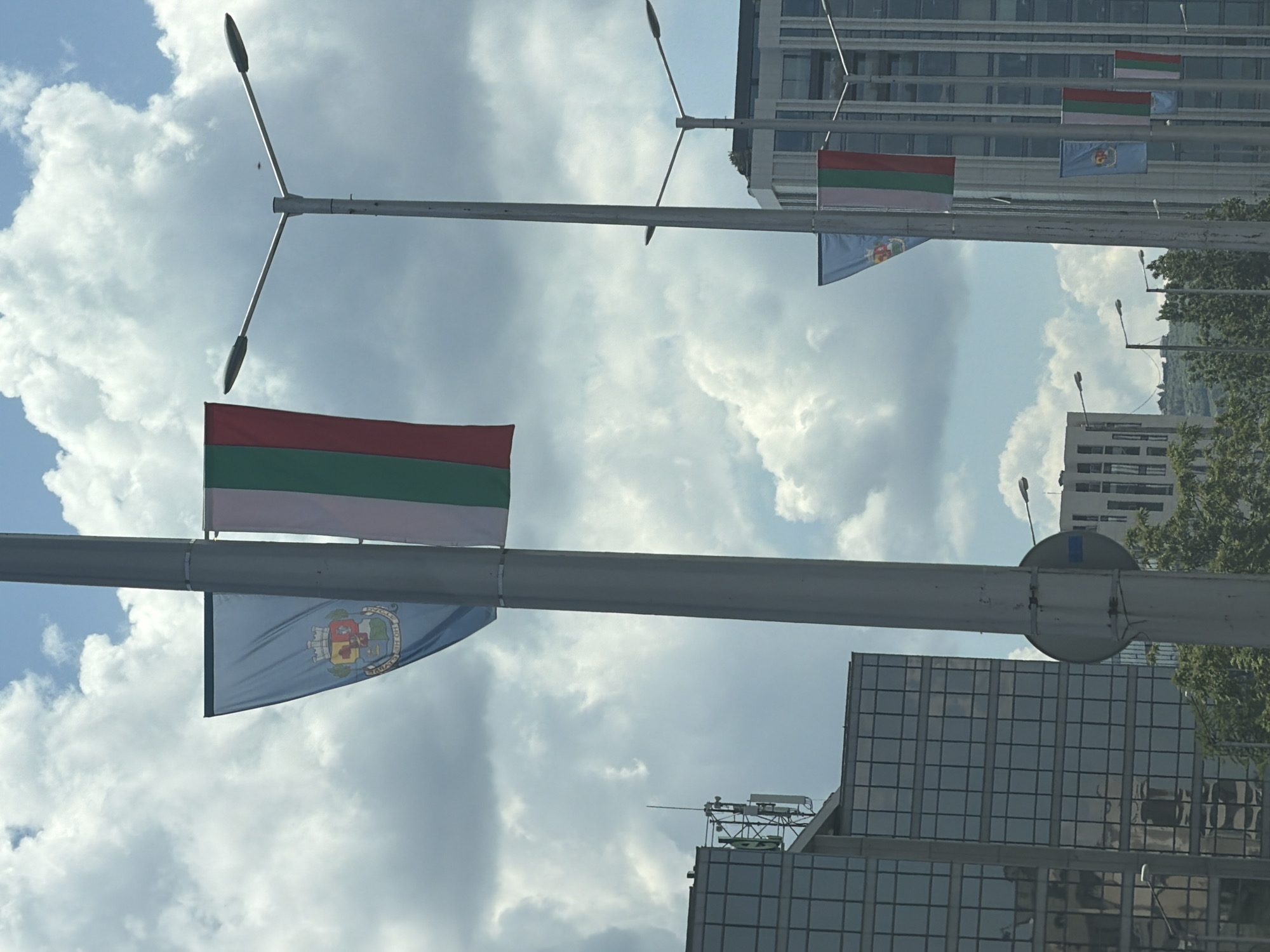 In the city of Sofia, Bulgaria, a street scene unfolds under a cloudy sky. A tall, gray lamppost dominates the foreground, from which hang two flags. The top flag, on the left, is light blue with a coat of arms featuring a crowned shield with various symbols and the inscription "СОФИЯ" in a curved line below. To its right, a tricolor flag with vertical stripes of red, white, and green is displayed. Further down the street, another lamppost supports a similar arrangement of flags. In the background, modern buildings with glass facades rise, interspersed with trees and more distant structures. The overall impression is one of an urban environment, possibly a governmental or administrative area, decorated with national and municipal symbols.