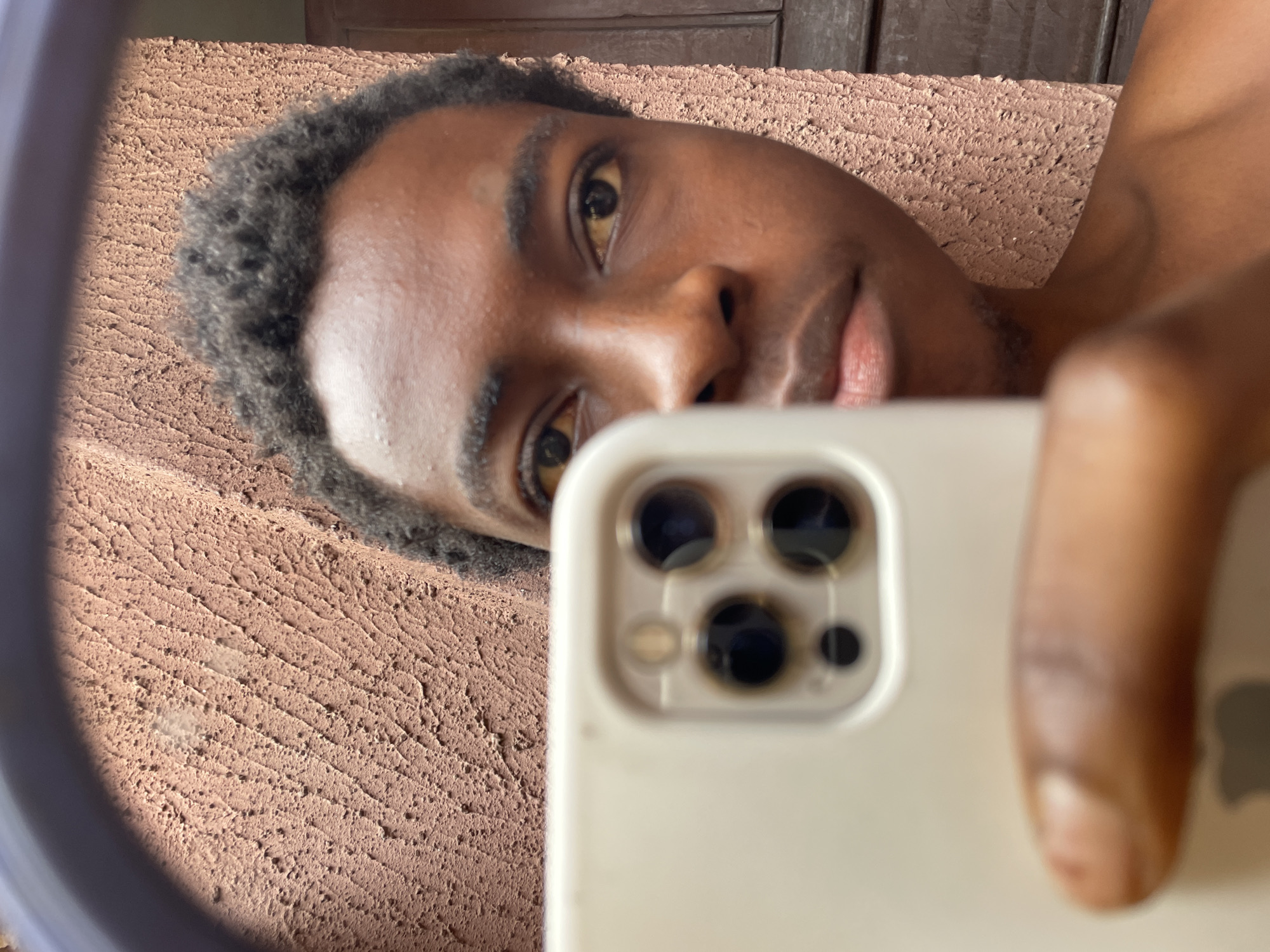 The image shows a selfie of a Black man with short, dark hair. He is holding a white iPhone with a triple-lens camera system. The background appears to be a textured wall, possibly of a building. The overall setting seems to be indoors. The location given, Nkpor, Nigeria, provides context for the environment and the subject's potential identity and origins. There are wooden panels above the textured wall.