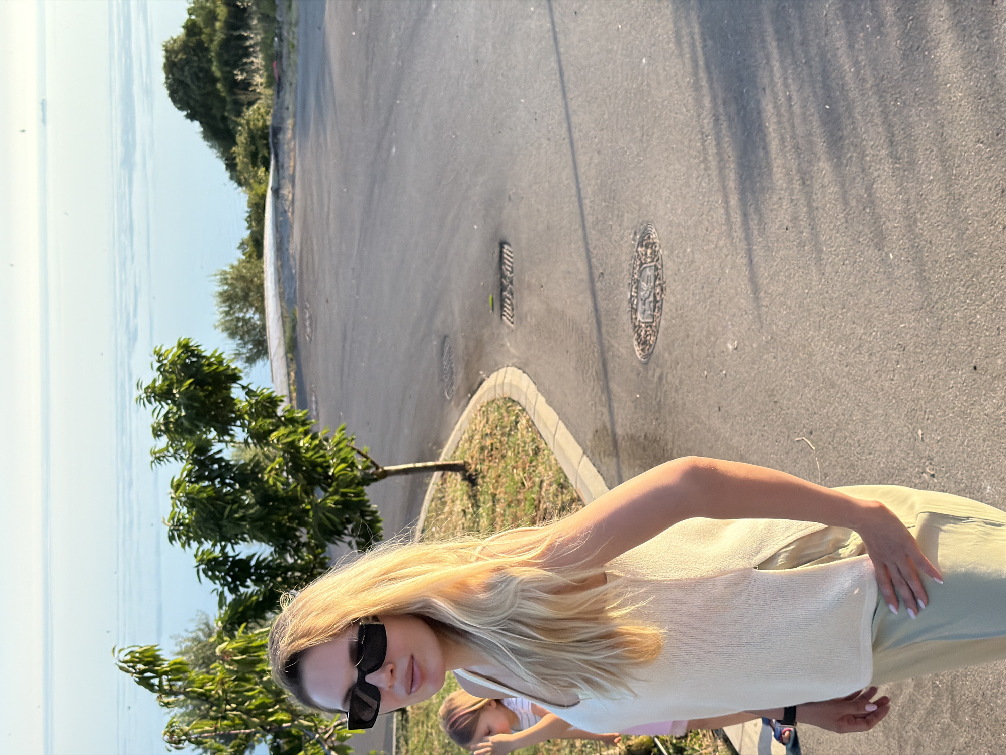 A woman with blonde hair and black sunglasses stands on a paved surface, looking to her left. She wears a cream-colored sleeveless top and light green pants. Behind her, a young child with blonde hair is partially visible, wearing a white and pink top. The background features trees, a body of water, and a distant shoreline. The scene appears to be outdoors, likely in a coastal area. The location is Kovačko Polje, Montenegro.
