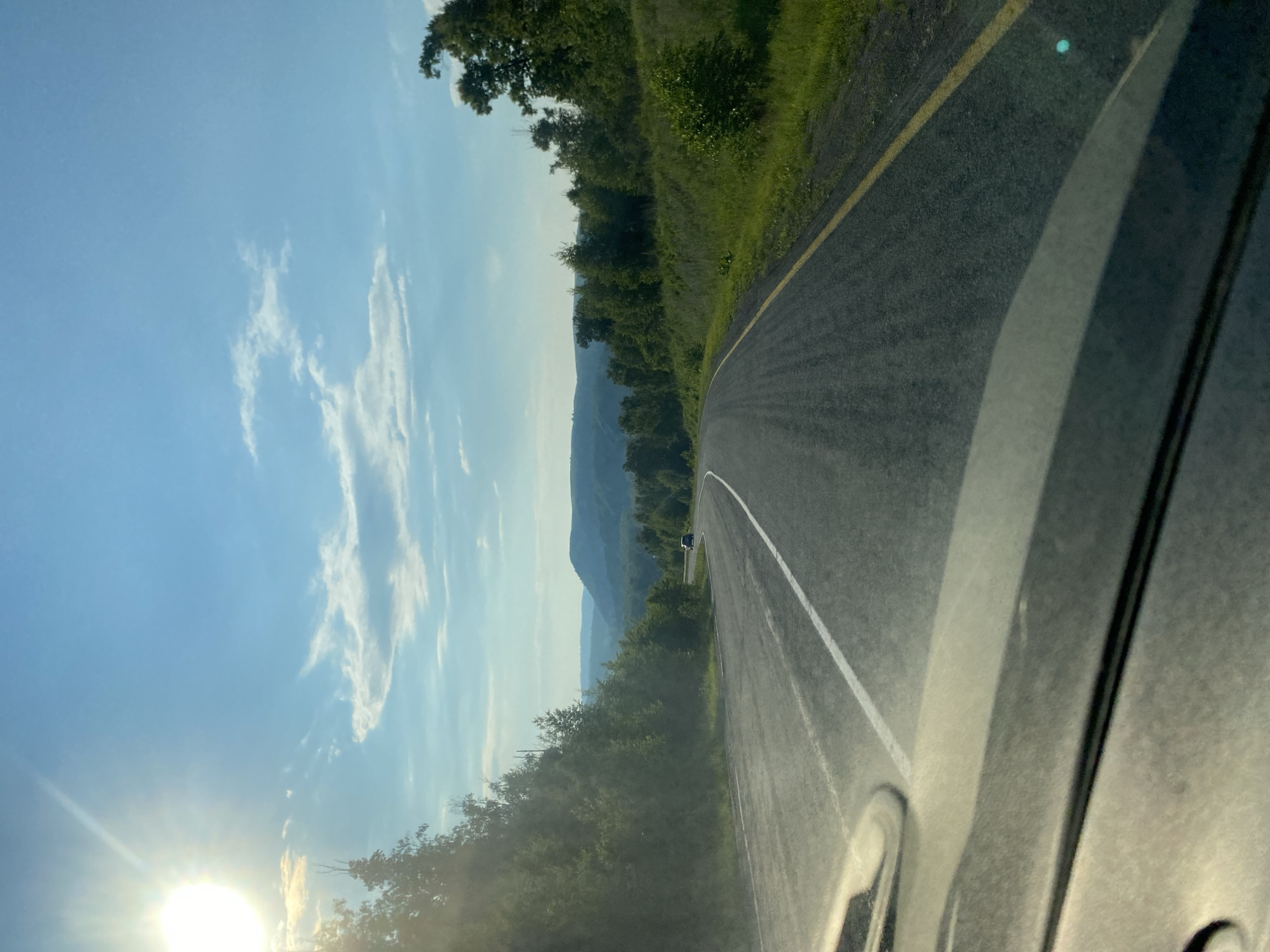 The image depicts a road winding through a hilly, forested landscape under a bright, sunny sky with scattered clouds. The sun is prominently visible in the upper left corner, casting a lens flare across the scene. The road appears to be paved with asphalt and has white and yellow lane markings. Trees and vegetation line both sides of the road, with a dense forest on the left and a mix of trees and grassy slopes on the right. In the distance, layers of rolling hills recede into a hazy blue horizon. A dark-colored car is visible in the distance, traveling in the same direction as the camera. The foreground shows the reflection on what appears to be a car's dashboard or windshield. The overall impression is a scenic drive through a rural or natural area. The specific location in the city of Антинган, Russia, cannot be determined from the visual information provided.