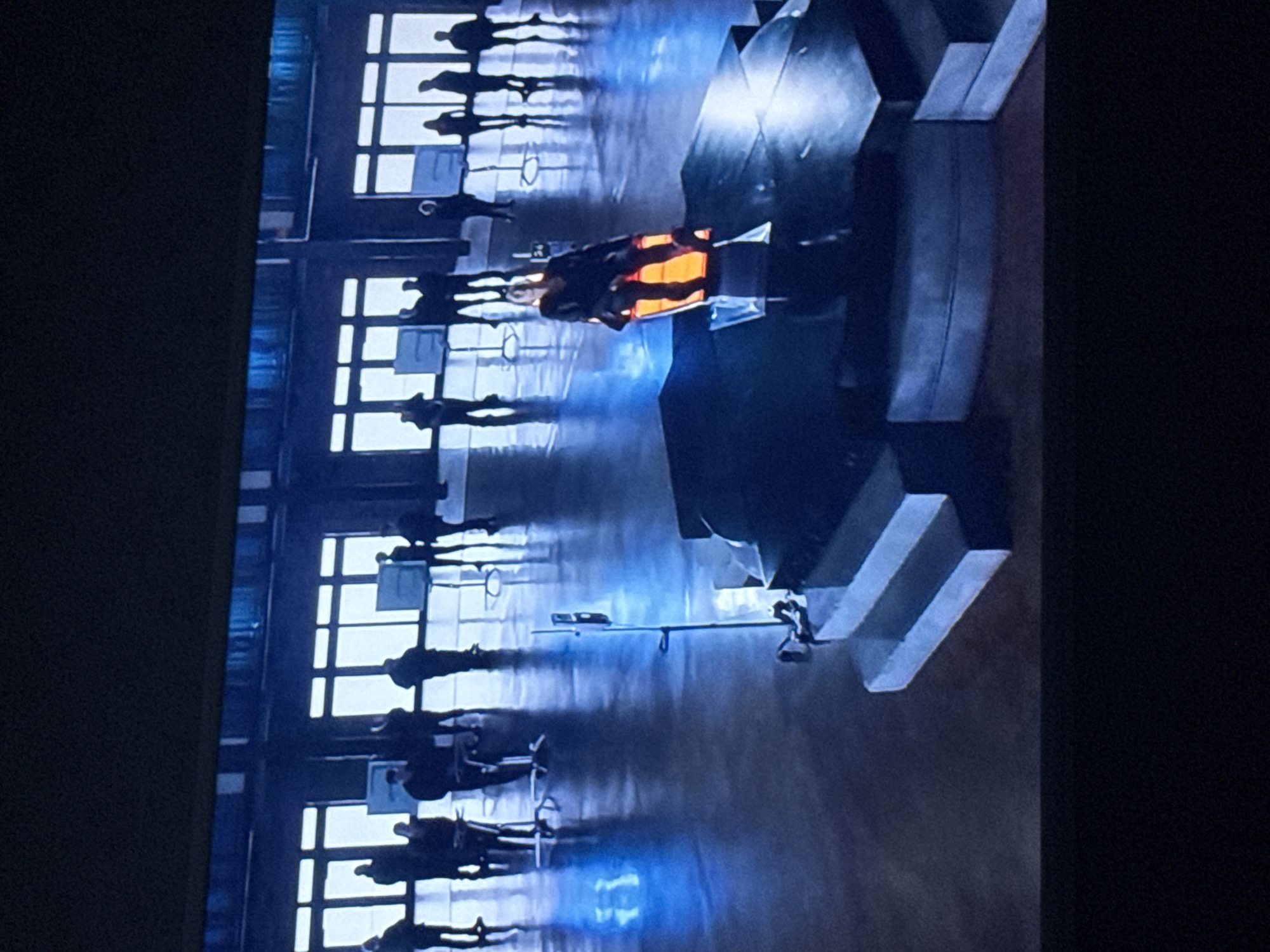 The image shows an interior scene. In the foreground, a large, dark structure with a semi-circular base and steps is visible. To the left, there are multiple figures lined up in rows, silhouetted against what appears to be a wall of windows with a grid pattern. The figures are standing at desks or tables, and there are hints of objects on these surfaces. The lighting appears to be blueish and somewhat dim. The environment is clean and institutional. There are no indications about a location in Mytishchi, Russia.