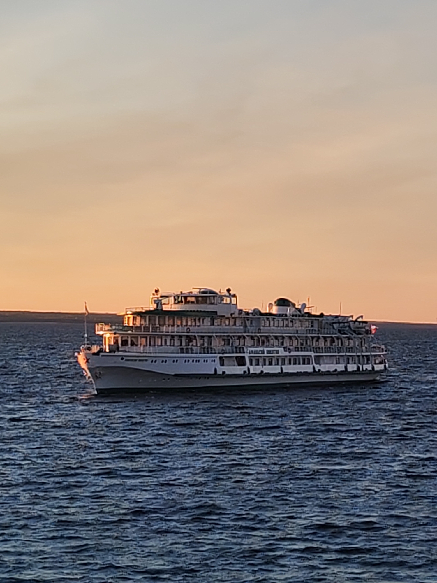 A large, multi-decked river cruise ship is traveling on water, possibly the Volga River, in Mikhalevo, Russia. The ship is white with dark trim and multiple rows of windows. The background features a sunset sky with colors ranging from orange to light grey. The water is dark blue and choppy. Land is visible on the horizon. No people are clearly visible.