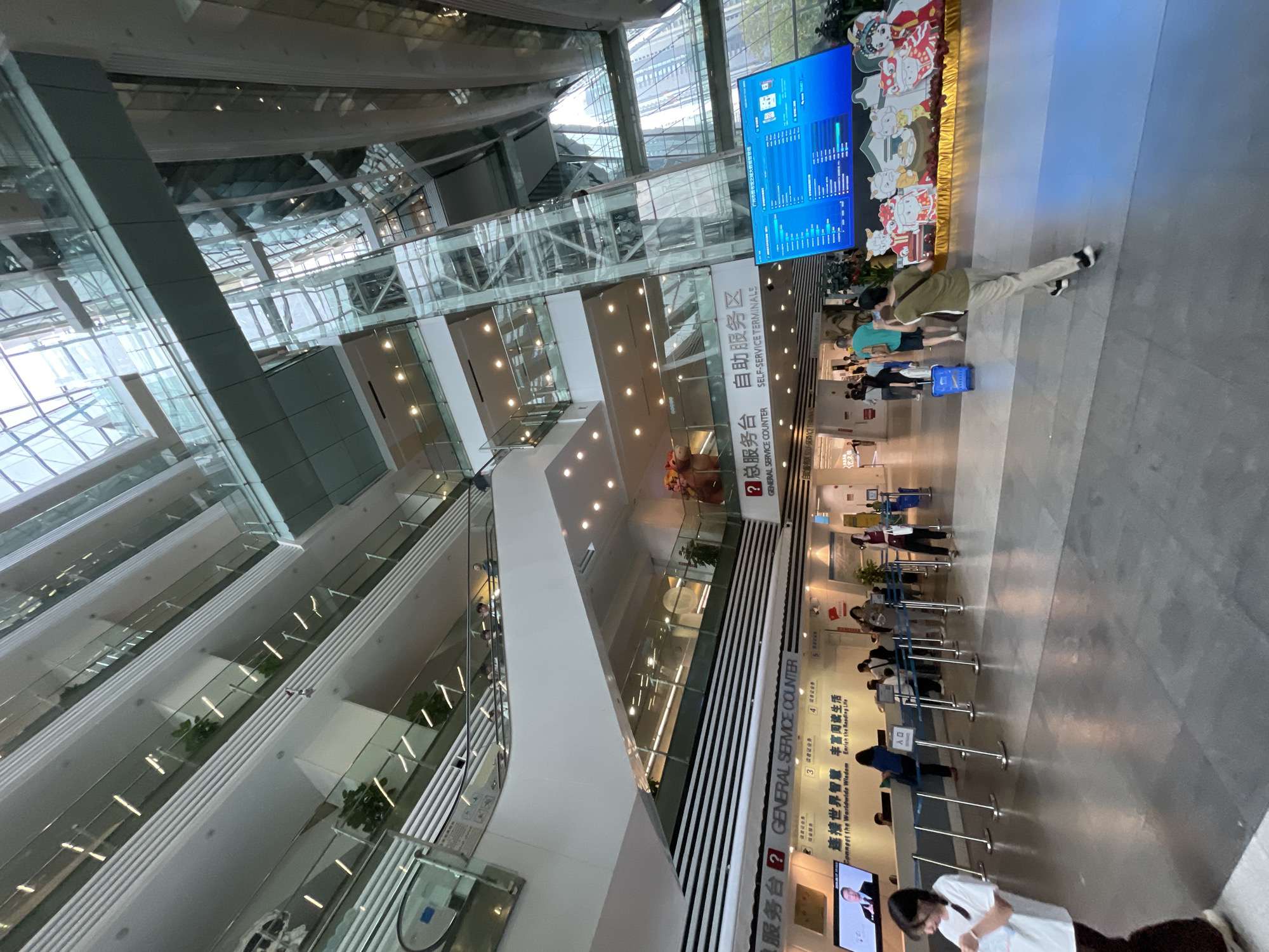 The image shows the interior of a multi-story building, likely a commercial or institutional space, in Tianhe District, China, specifically its General Service Counter. The architecture features a modern design with glass panels and internal balconies adorned with plants. A large digital display board is visible near the top of the frame, alongside decorative elements. There are several people present, some standing in lines at service counters, while others are walking through the space. The overall lighting is bright and artificial. Banners with Chinese characters are prominently displayed, including the words "General Service Counter," and "Self-Service Terminals."