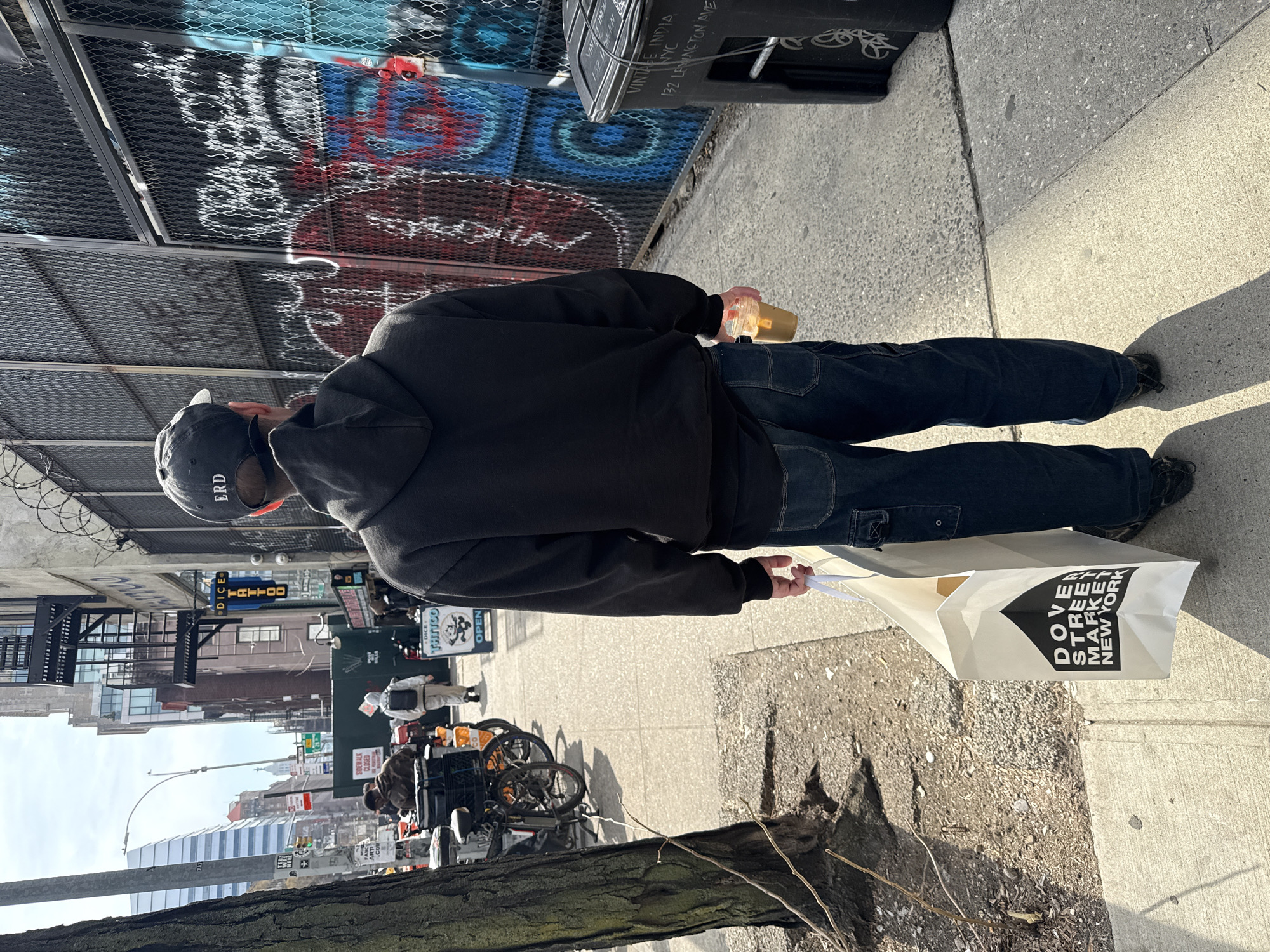 A male individual is depicted from behind, walking southbound on a sunlit sidewalk on Lexington Avenue in New York, United States of America. He wears a black hooded sweatshirt, dark blue jeans, and a black baseball cap with "ERD" embroidered in white on the back. His left hand holds a white shopping bag displaying "DOVER STREET MARKET NEW YORK," while his right hand holds a clear plastic cup containing a light brown beverage.
To the individual's right, a chain-link fence topped with barbed wire is extensively covered in multi-colored graffiti. Adjacent to the fence is a black plastic trash receptacle with "VINTAGE INDIA NYC 132 LEXINGTON AVE" stenciled in white. To the individual's left, a large tree trunk borders the sidewalk.
Further down the street, multiple pedestrians are visible, along with several parked bicycles, some appearing to be delivery bikes equipped with large carriers. A green construction barrier with a "SIDEWALK CLOSED" sign partially obstructs the view. A blue neon sign for "DICE TATTOO" is mounted on a building, with an additional "DICE TATTOO OPEN" sign positioned on the sidewalk below it. Various urban buildings, ranging in height and architectural style, line the street under a clear, bright sky.