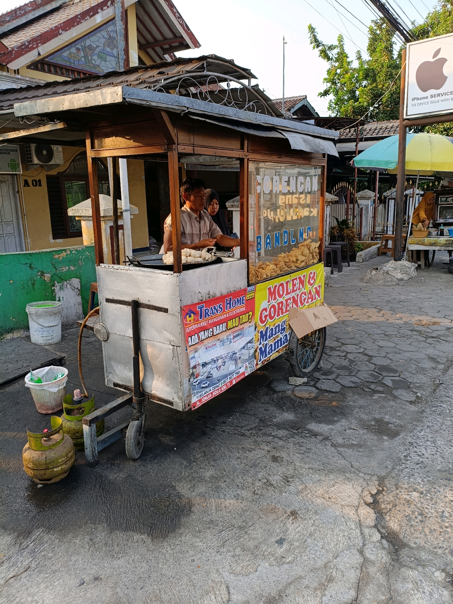 This is a medium shot of a food cart vendor in Purwomartani, Indonesia. The cart, made of wood and metal, is on wheels and appears to be a mobile food business. The vendor, a man wearing a striped shirt, is operating the cart. He is standing behind a display of fried food, possibly "gorengan" (Indonesian fritters), in a glass enclosure. A woman in a headscarf is also present, near the vendor. Two gas cylinders are visible on the left side of the cart, one upright and one tilted. The cart has signage with text in Indonesian, including "MOLEN & GORENGAN" and "Mang Mama," indicating the food being sold. In the background, there's a building with an air conditioning unit, a stained-glass window on its facade, and some trees. A sign with an Apple logo and "iPhone Service" is visible on the right. The ground is paved with cobblestones. The lighting suggests it's daytime, with sunlight casting shadows.