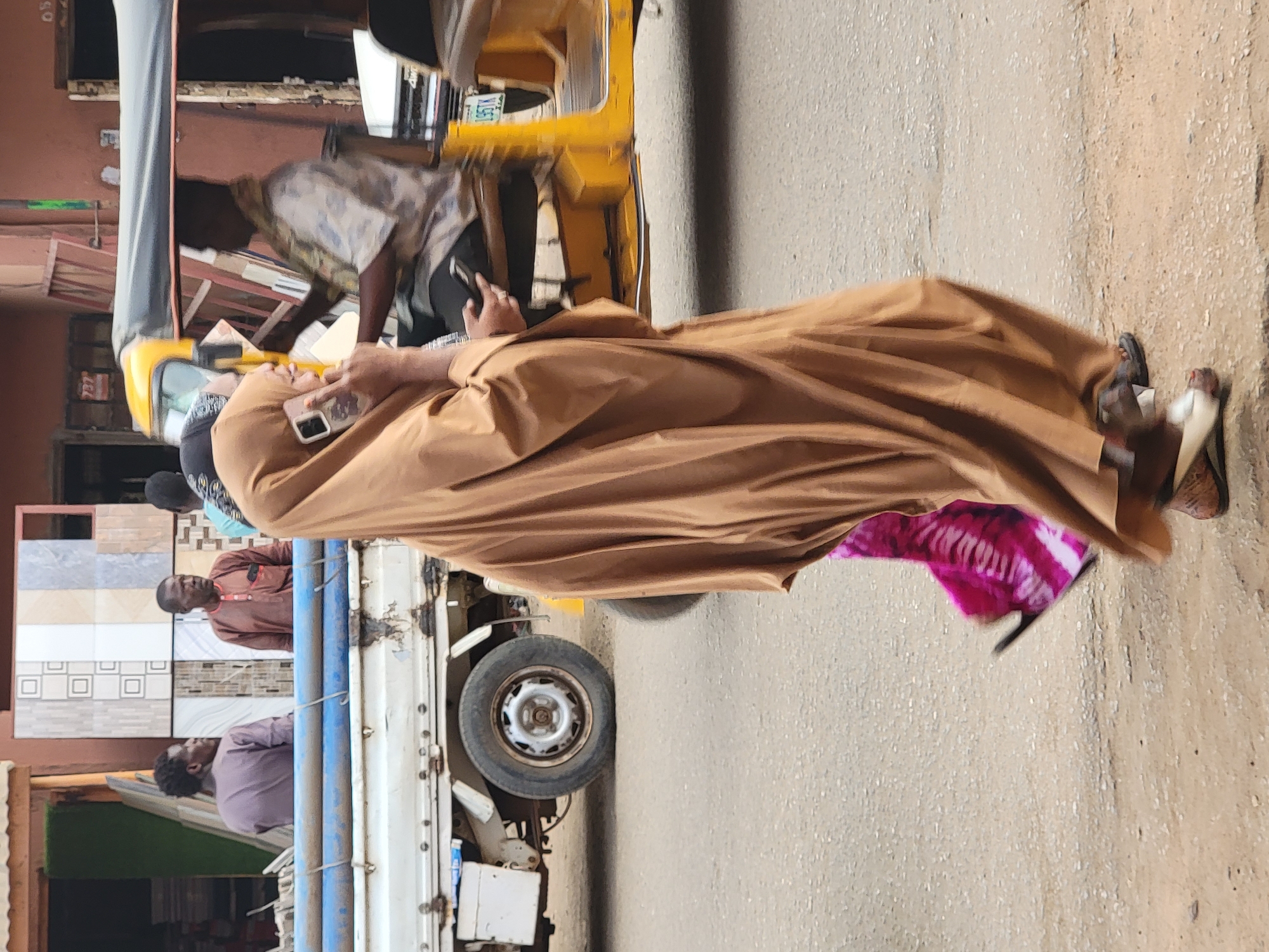 The image shows a street scene in Maiburiji, Nigeria. A man wearing a brown robe is walking on the paved street, looking at his phone. A yellow tricycle taxi is parked to the left of the walking man. A truck is loaded with tiles. People stand near the truck. The buildings are visible in the background.