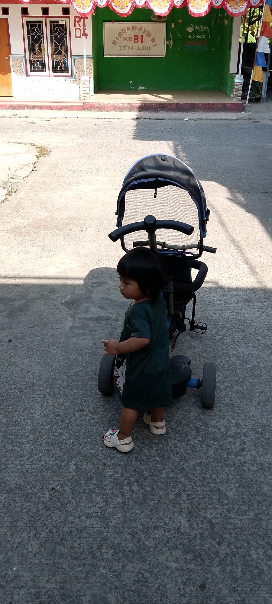 The image shows a young child, appearing to be a toddler, standing on a paved surface next to a blue and black stroller. The child is wearing a dark green dress and light-colored sandals. The child is facing away from the camera and appears to be looking to the left. The stroller is a three-wheeled model with a fabric canopy and a handlebar. In the background, there is a building with a white facade and red trim, and a green wall with a framed sign that reads "DIRGAHAYU KE 81 RT 04 AKUR". The building appears to be part of a residential or community area in Satriamekar, Indonesia, possibly a Posyandu or community center, given the sign. The ground is paved with concrete and shows signs of wear. The lighting suggests it is daytime with direct sunlight casting shadows.