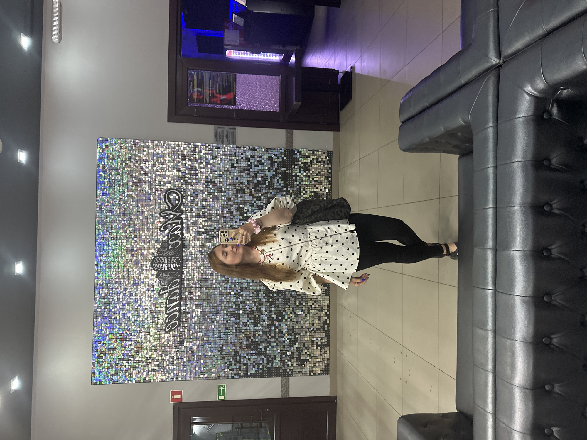 A woman with long, reddish-brown hair is taking a selfie in a brightly lit, modern-looking establishment. She is wearing a white blouse with black polka dots and puff sleeves, black leggings, and black heeled sandals. She is also carrying a large, dark-colored patterned tote bag. Behind her is a large, glittering wall made of small, reflective squares, with a stylized cityscape and text in black. To her left is a closed door with a fire extinguisher sign. To her right is another doorway leading to a dimly lit room with blue lighting and what appear to be arcade or entertainment machines. In the foreground, a section of a black leather tufted sofa is visible. The floor is tiled. The overall impression is that of an entrance or lobby area of an entertainment venue.