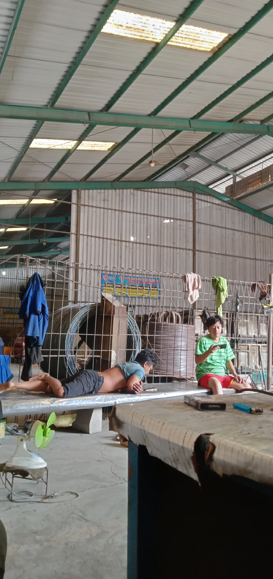The image shows an indoor scene in Banjaran, Indonesia, likely a workshop or industrial space. Two men are present. One is lying prone on a flat surface, possibly a metal platform, looking at a mobile phone. He is wearing shorts and a t-shirt. The other man is seated nearby, wearing a green shirt and red shorts, also looking at a mobile phone. A pack of cigarettes and a lighter are visible on a table near him. The background includes a wire cage containing coils of wire, a blue jacket hanging on the cage, and a corrugated metal wall. The space has a high ceiling with visible support beams. A small electric fan sits on a stand near the man lying down. The floor appears to be concrete.