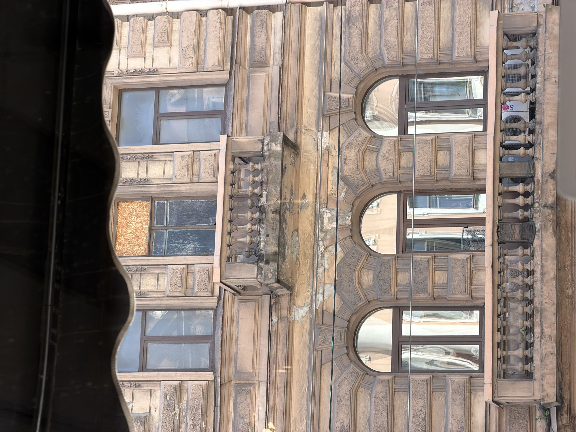 The image shows the facade of a building in Saint Petersburg, Russia, with several windows. The architecture appears classical with decorative elements. Some windows are rectangular, while others are round. One window is boarded up. There's also a decorative column-like structure on the right side of the frame. The photo seems to have been taken from inside a building, looking out through a window frame.