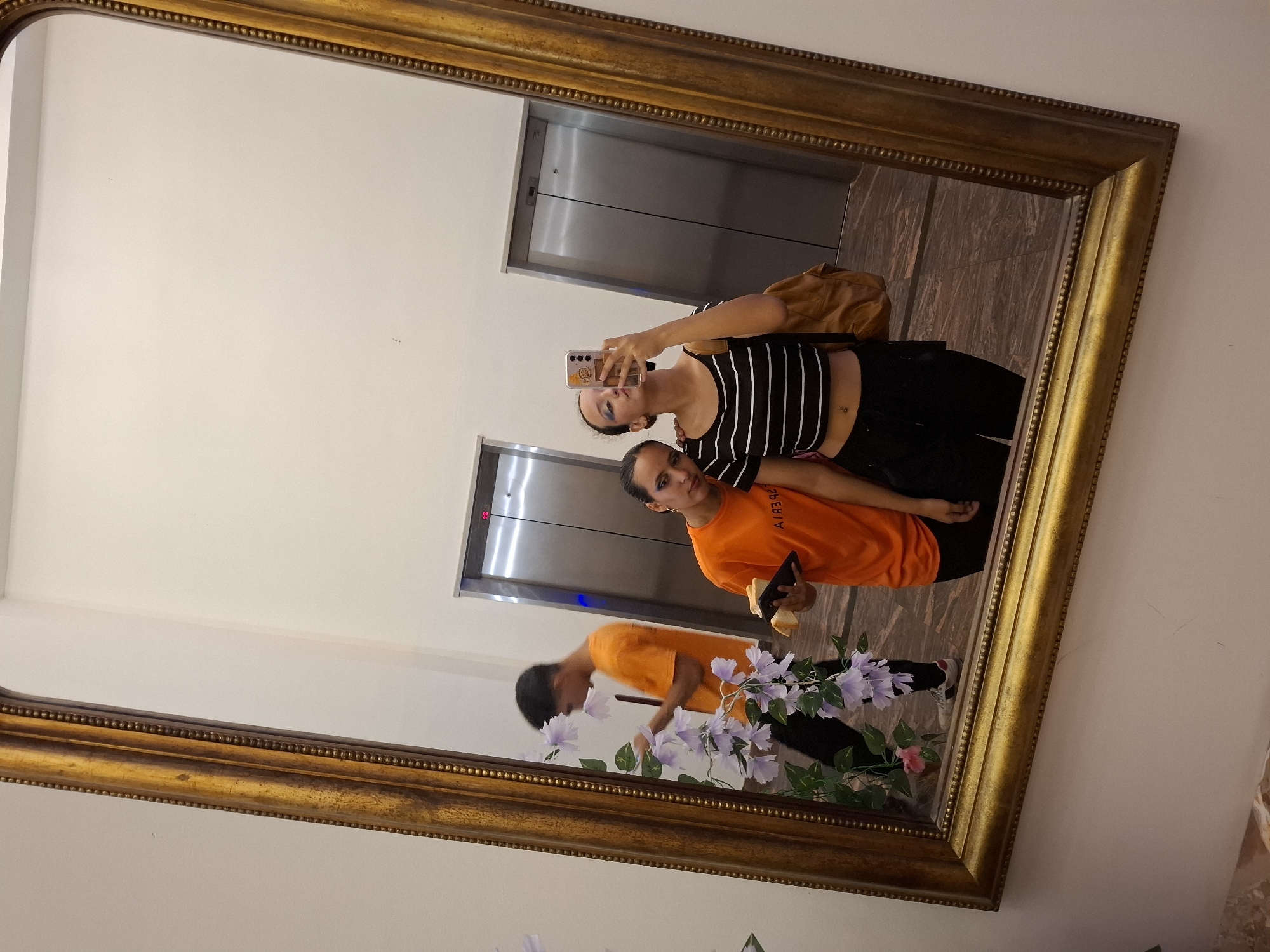 A tilted, rectangular, ornate gold-colored framed mirror hangs on a light beige wall, reflecting a scene. In the reflection, two individuals are present. One is holding a phone, presumably taking the picture, wearing a black and white striped top and black pants, with a brown bag slung over the shoulder. The other is wearing an orange shirt and black pants, holding what appears to be a pastry. They appear to be posing or taking a selfie together.
Behind them in the reflection is an elevator bank. A flower arrangement is also visible in the reflection, positioned at the bottom of the frame.
While it is impossible to confirm the location without additional information, the prompt suggests it is in Pedro González, Venezuela. However, there is no way to verify this solely from the image.