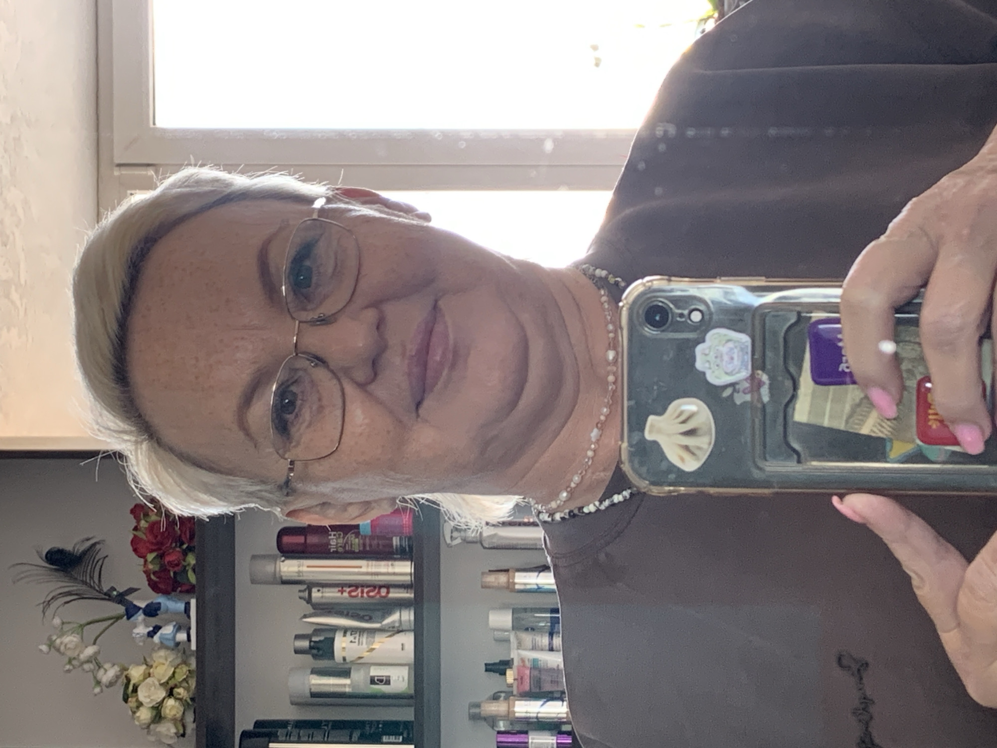 A person with fair skin, wearing glasses and a beaded necklace, is holding a smartphone in front of their face, likely taking a selfie. The smartphone case has stickers on it. The person is wearing a brown top. Behind the person, a shelf is visible, containing various cosmetic products, tubes, and cans. There's also a small floral arrangement with a peacock feather on the shelf. Based on the presence of products in Russian language, there is a possibility that the location is the city of Chaikovsky, Russia.