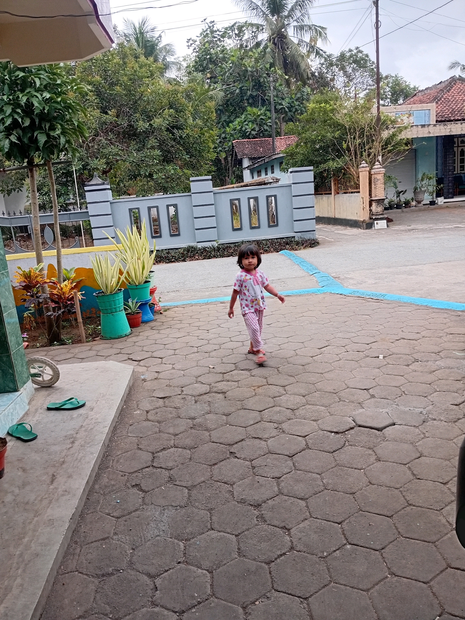 In Gebang, Indonesia, a young girl with dark hair is walking on a hexagonal paved courtyard. She is wearing a pink and white polka dot shirt and pink striped pants, along with brown sandals. The courtyard is bordered by a gray wall with rectangular window-like openings and decorated with potted plants. To the left of the courtyard, there is a concrete step with green flip-flops and a white wheel. Beyond the wall, trees and houses are visible under an overcast sky.