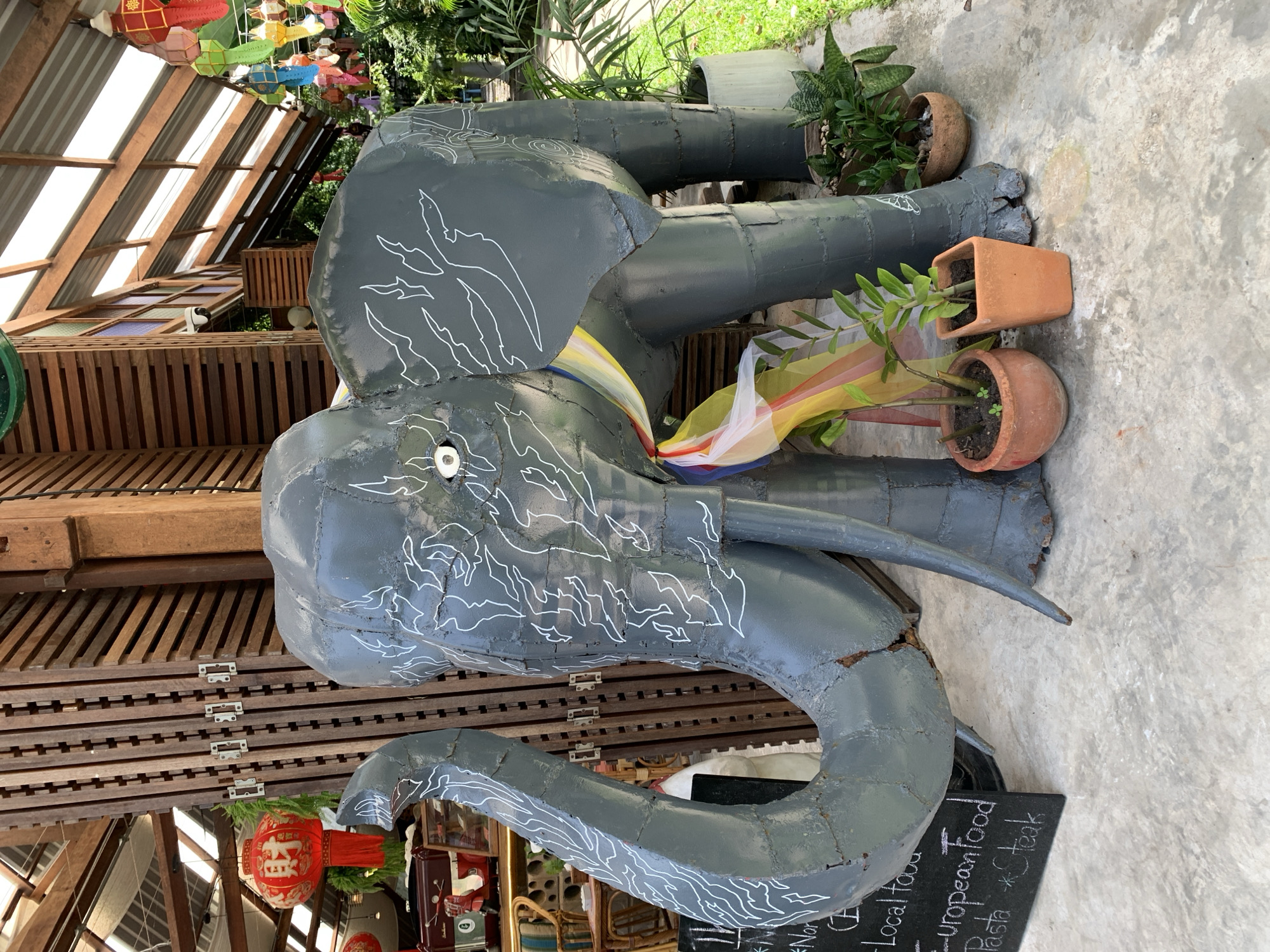 An outdoor scene depicts a grey metal elephant sculpture adorned with white swirling patterns, prominently positioned on a concrete floor. The sculpture is accessorized with a draped colorful ribbon around its neck. To the left of the elephant, a black chalkboard displays menu items including "Local food," "European food," "Pasta," and "Steak." Behind the sculpture, a wooden wall with vertical slats is visible, and hanging above are red and gold Chinese lanterns, suggesting a festive or cultural setting. The background reveals a covered area with a transparent roof, sunlight filtering through, and lush green foliage, indicating an outdoor dining or market environment. The scene appears to be located in the city of Ko Samui District, Thailand, given the cultural elements and tropical vegetation.