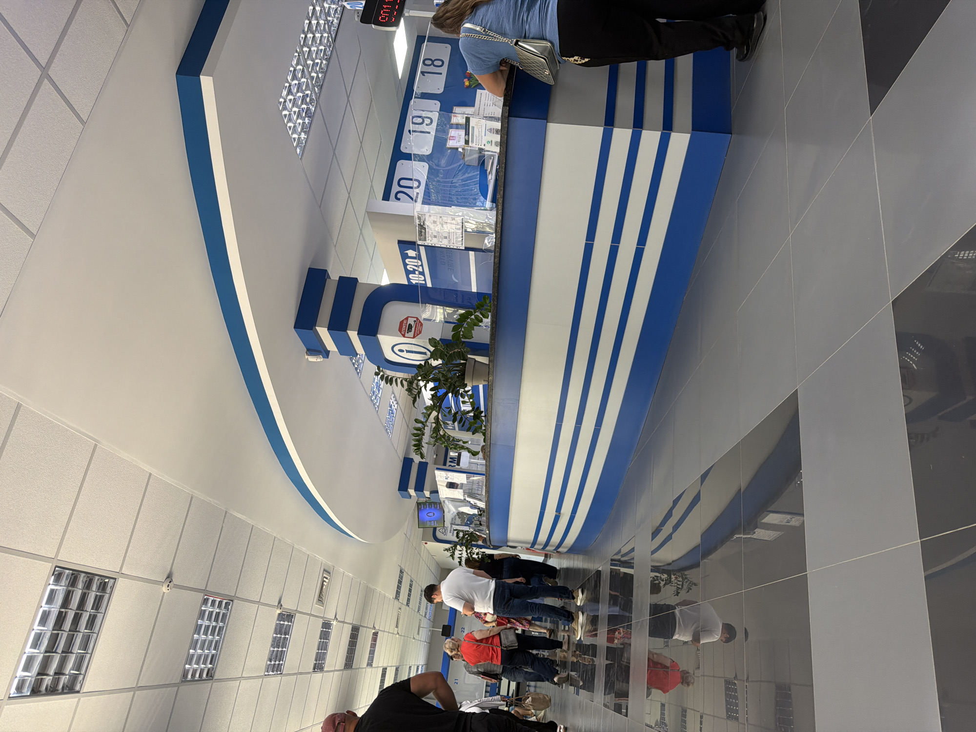 The image depicts the interior of a modern, well-lit service center in Strășeni, Moldova. The facility features a long, curved counter with blue and white stripes, indicating a professional and clean aesthetic. Fluorescent ceiling lights are visible, and the floor is tiled with reflective grey and black squares, which create reflections of the people and objects in the room.
Several individuals are present, suggesting a queue or service area. In the foreground to the left, a person wearing a black t-shirt is partially visible from behind. Further into the scene, a group of people stand near the counter, including a woman in a red shirt, a man in a white t-shirt, and another woman. They appear to be waiting or being served. On the right side of the counter, a woman with long brown hair, wearing a blue shirt and dark pants, is interacting with someone behind the counter.
The counter itself has clear partitions, information displays, and a potted plant, contributing to a functional and organized environment. Numbered signs, such as "20", "19", and "18", are visible above the counter, possibly indicating service stations or queues. A digital display showing "001" is also present, likely related to the queue management system. The overall impression is of an administrative or public service building.