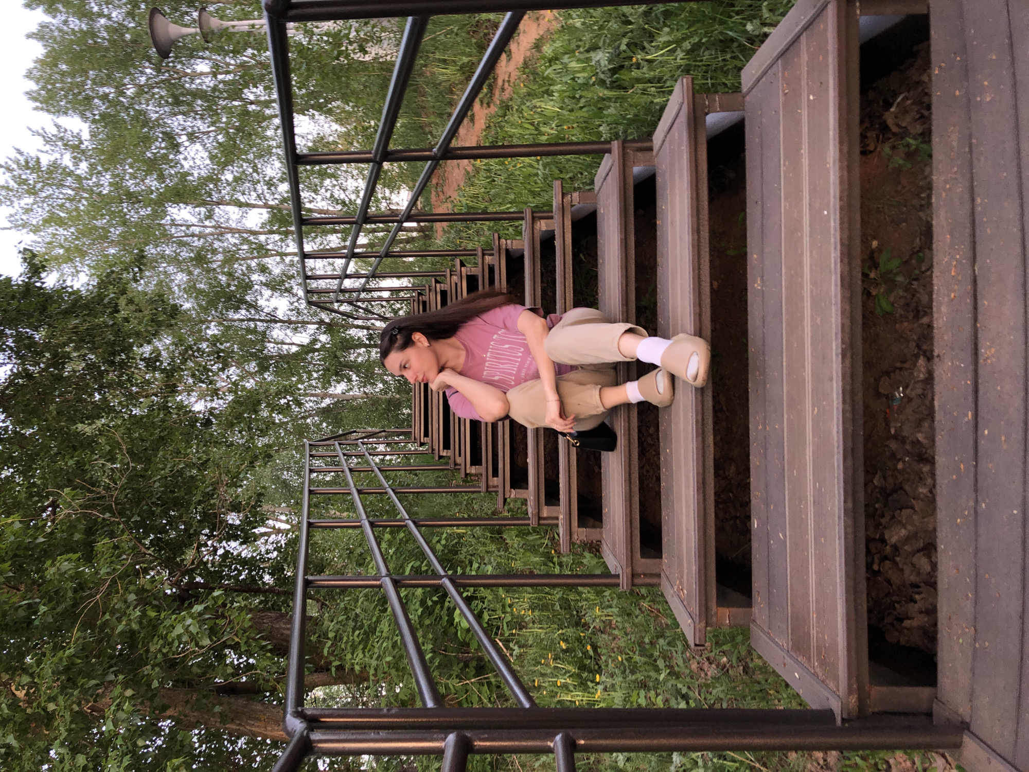 A woman is sitting on a wooden staircase, which is likely located in the city of Chaikovsky, Russia. The staircase features a metal railing on either side. The woman is wearing a pink t-shirt, khaki pants, and tan slippers with white socks. She is posed thoughtfully with her head resting on her hand. The area surrounding the staircase is green with trees and vegetation.