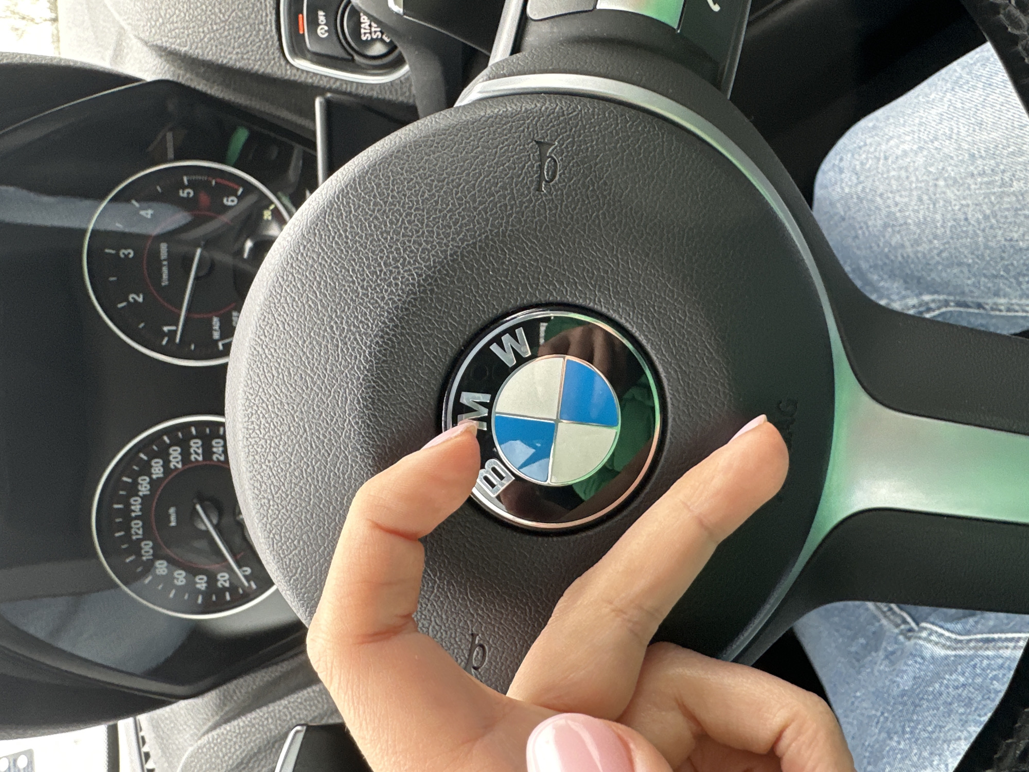 A close-up shot inside a vehicle, likely a BMW, focuses on the steering wheel. A hand with light pink nail polish is positioned in front of the BMW logo on the steering wheel. The dashboard displays the speedometer and tachometer. The speed indicated on the speedometer is difficult to discern. The car appears to be in an urban environment, although the specific city (Sofia, Bulgaria) cannot be determined from the image alone.