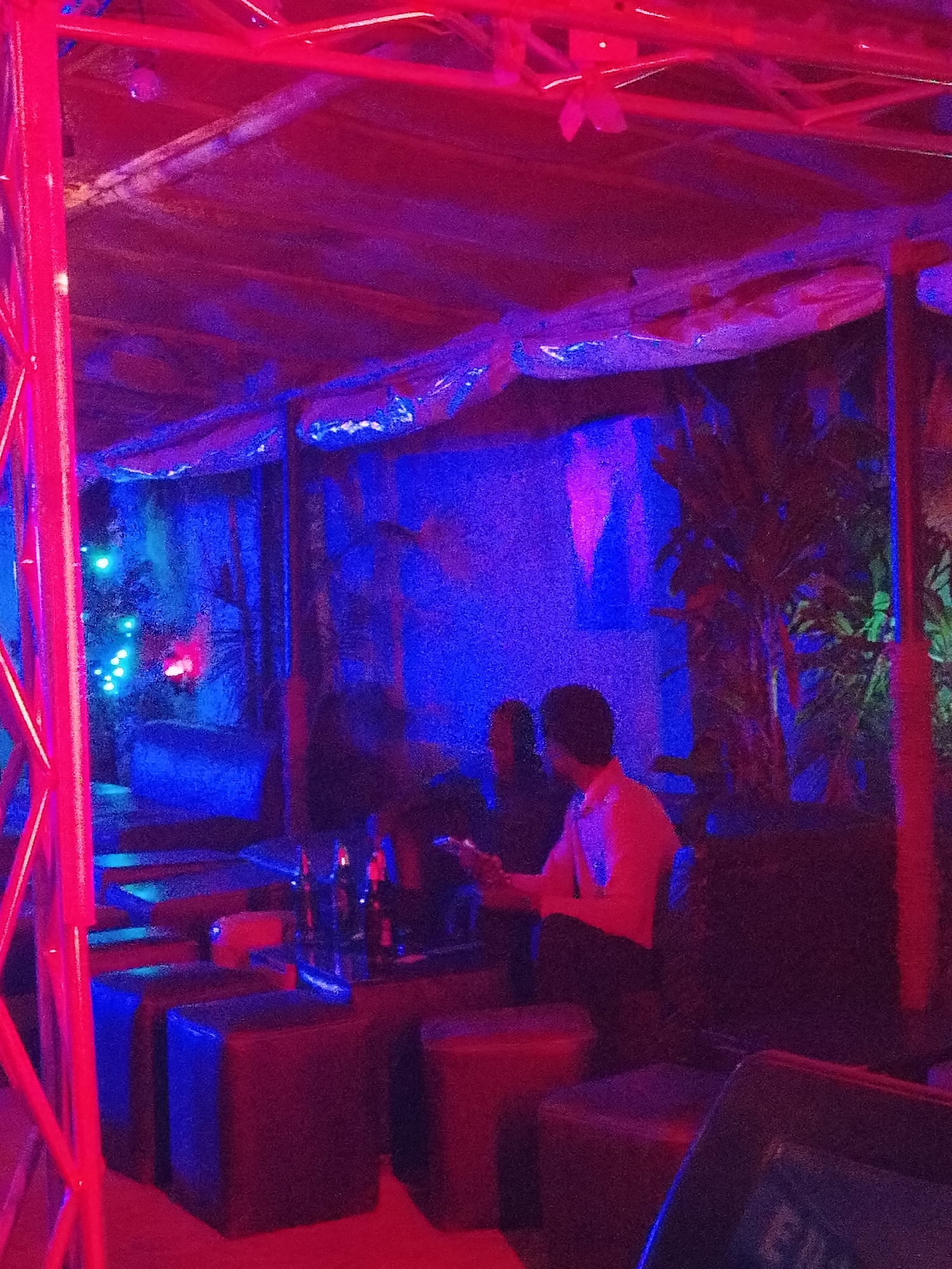 Here is a description of the image:
The image shows the interior of a bar or lounge in the city of Unknown, Unknown, illuminated by blue and red neon lights. The foreground features a red scaffolding structure. A small table with several bottles on it is visible near the center. Two people are seated at the table. The person on the right is wearing a light-colored shirt and dark pants, and appears to be looking at a device. The second person is mostly obscured in the dark. The seating consists of cushioned, dark-colored stools or small ottomans. The background includes an arrangement of foliage. The overall environment has a dark and moody atmosphere.