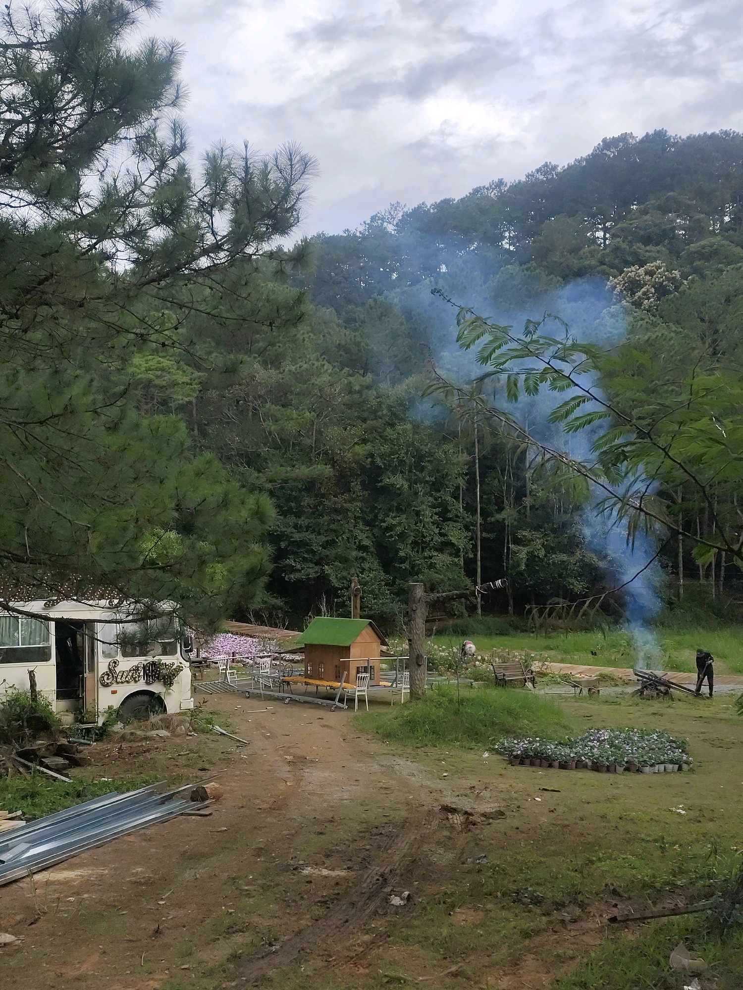 The image depicts an outdoor scene in the city of Da Lat, Vietnam, likely a rural or semi-rural area given the abundance of trees and foliage. In the foreground, a dirt path leads into a clearing. To the left, a beige bus with the name "Sankey" visible on its side is parked. The bus appears to be modified, potentially for living or as part of an establishment. In the clearing, a small, green-roofed wooden structure resembling a playhouse or miniature building is situated on a table. Various outdoor seating arrangements, including white chairs and a wooden bench, are scattered around the area, interspersed with potted plants and a patch of pink flowers.
Further into the background, a dense forest of pine and other trees covers the sloping terrain, characteristic of Da Lat's mountainous landscape. A plume of blue smoke rises from a fire pit in the mid-ground, where a person dressed in dark clothing is tending to the flames. This suggests an ongoing outdoor activity, possibly cooking or clearing debris. The sky is overcast with a mix of light and dark clouds, indicating a potentially cool or damp day. The overall impression is one of a rustic, outdoor establishment or campsite nestled within a natural, wooded environment.