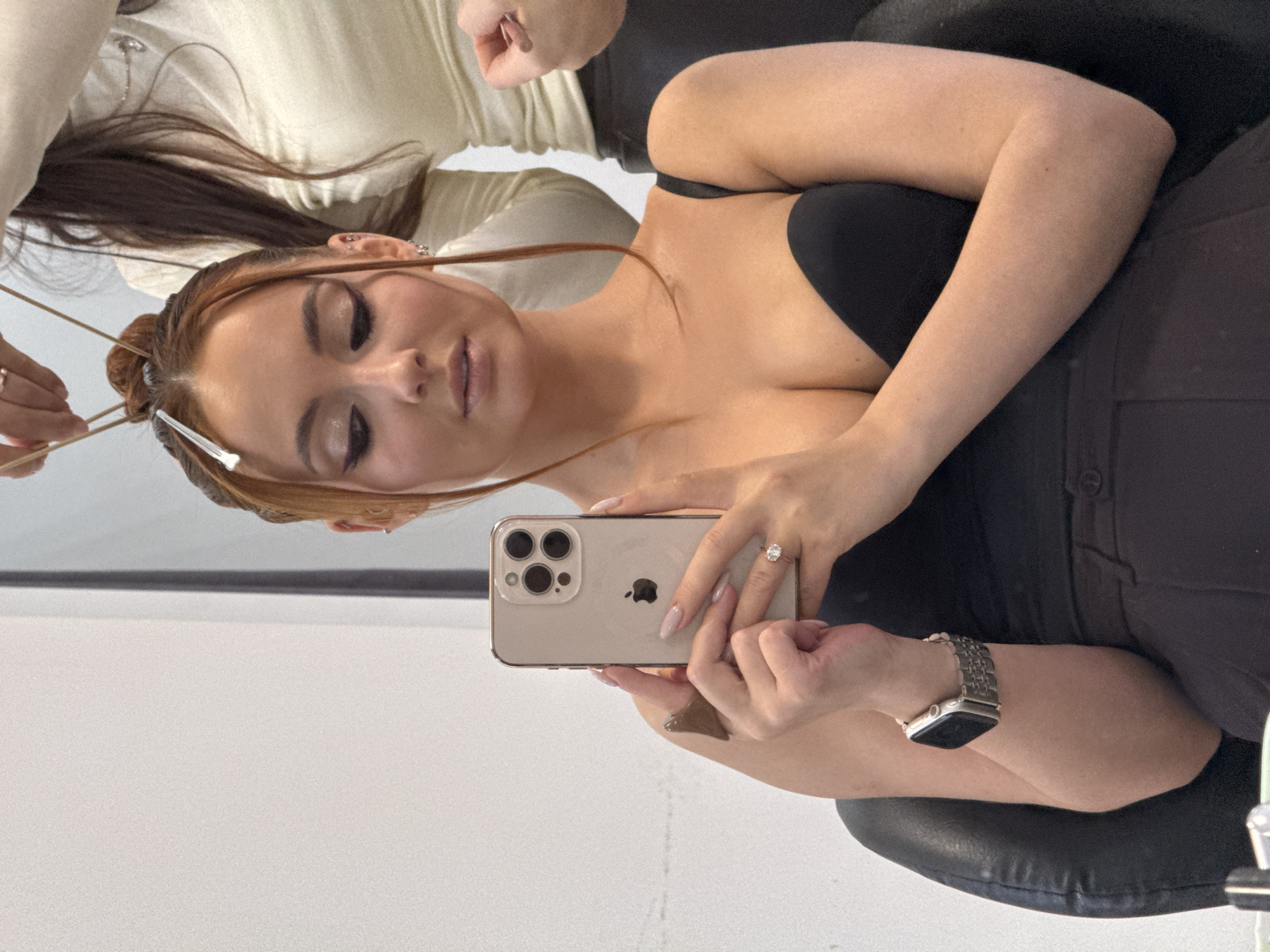 The scene is an interior space, likely a hair salon or beauty studio, within the city of Chaikovsky, Russia; its specific address is not discernible from the image.
The image features two individuals. The primary subject is a woman, appearing to be in her 20s or 30s, seated in a black, padded chair. She has auburn hair styled into an updo, secured with a silver hair clip near her forehead and with strands framing her face. Her eyes are adorned with prominent winged eyeliner and eyeshadow. She wears a black strapless top and dark trousers. On her left wrist, she wears a silver-toned Apple Watch with a link bracelet. Her right hand, exhibiting light pink manicured nails, holds a light-colored iPhone (with a triple-lens camera array visible) vertically, angled as if taking a selfie or viewing her reflection. A ring with a clear, oval-shaped stone is visible on her right ring finger.
A second individual, a hairdresser or stylist, is partially visible behind and to the right of the primary subject. This individual's right hand is engaged in manipulating strands of the primary subject's hair with thin, dark elastic bands or strings, indicating a hairstyling action. This person wears a light-colored, long-sleeved top and dark trousers, and a silver necklace is visible on her neck.
The background consists of a plain, light-colored wall. The primary subject is looking downwards towards the iPhone screen, while the secondary individual focuses on the hair styling.