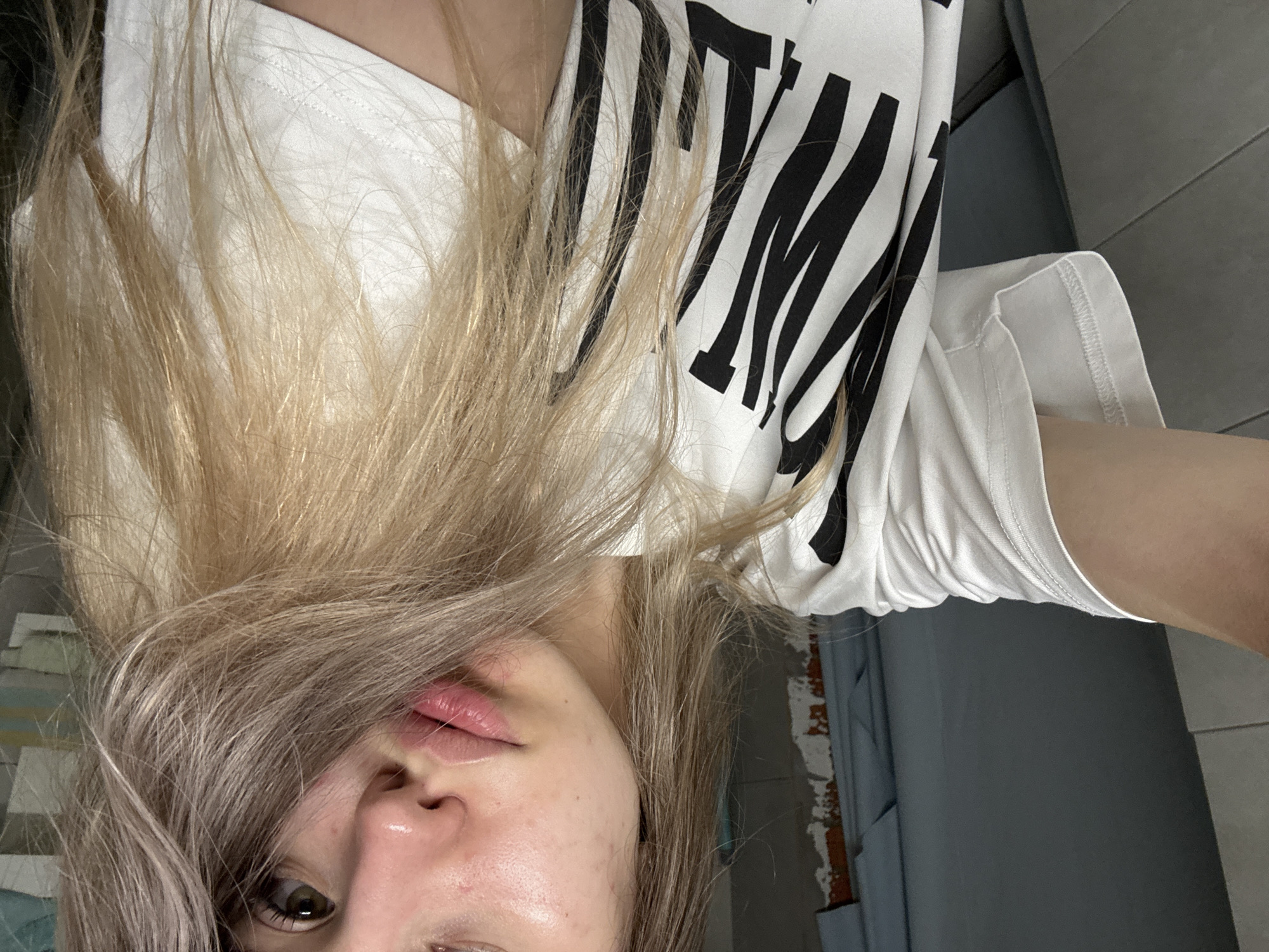 A selfie depicts a fair-skinned individual with blonde and gray hair, wearing a white t-shirt with black lettering, oriented upside down. The background shows a blue wall with a white, tiled section on the left and a gray wall on the right. The location is possibly Chaikovsky, Russia.