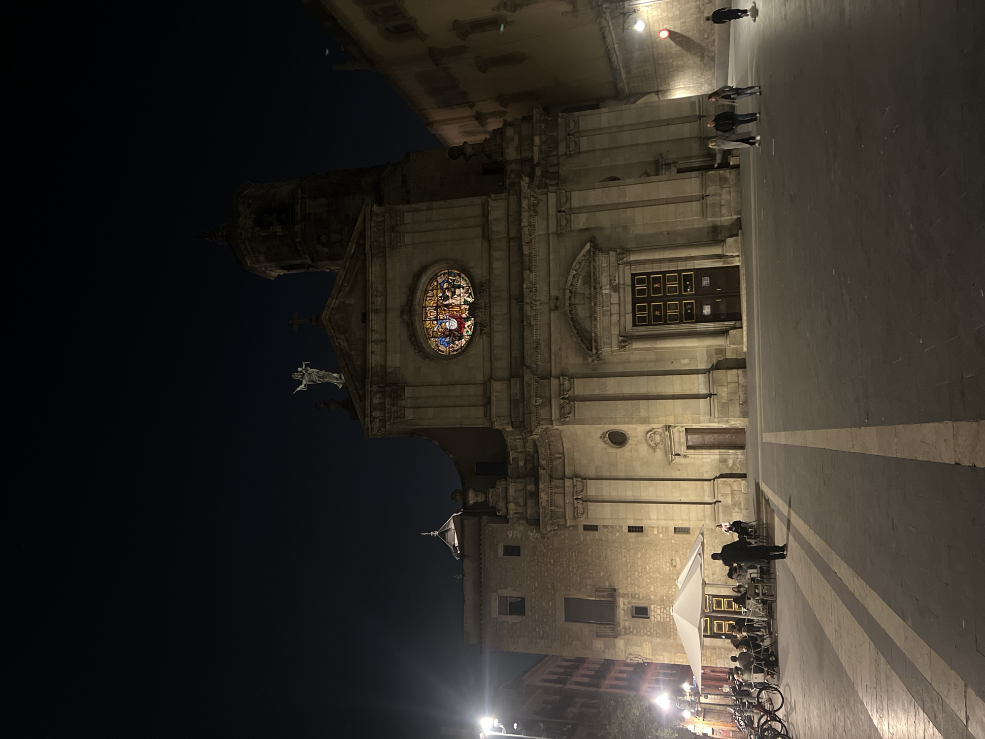 Captured at night, the image displays the impressive facade of a historic stone church or cathedral, likely in Barcelona given the context, dominating the left side of a spacious public square. The building, constructed of light-colored stone, features a large, illuminated circular stained-glass rose window adorned with vibrant patterns in its upper section. Above the rose window, a statue of a religious figure is visible on a ledge. Below the window, there is a grand wooden entrance door composed of numerous rectangular panels. To the left of the main facade, a tall, multi-tiered bell tower stands against the dark night sky.
The expansive paved square, stretching across the right side of the image, exhibits distinctive light and dark striped patterns. Several people are scattered throughout the square. In the foreground on the right, a small group of individuals is seen standing or walking. Further back on the right, an active outdoor cafe area is illuminated, with patrons seated at tables beneath a large white umbrella. Bicycles are parked near the cafe. Other older stone buildings with windows and balconies can be seen in the background behind the cafe.
The scene is set at night, with streetlights casting a warm glow on the architecture and the square. The overall impression is one of a calm and social evening in a historic European city center. No visible text is legible within the image.